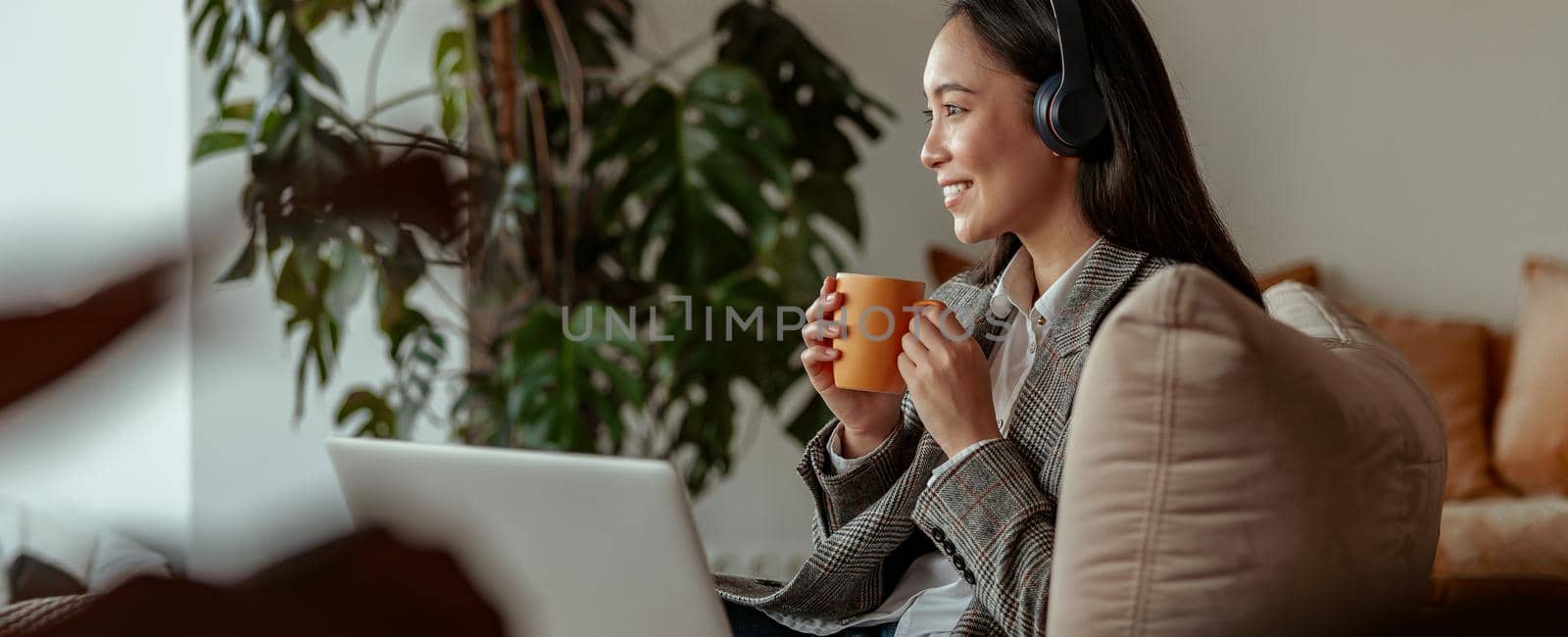 Attractive asian business woman listening music on headphones sitting at laptop with coffee by Yaroslav_astakhov