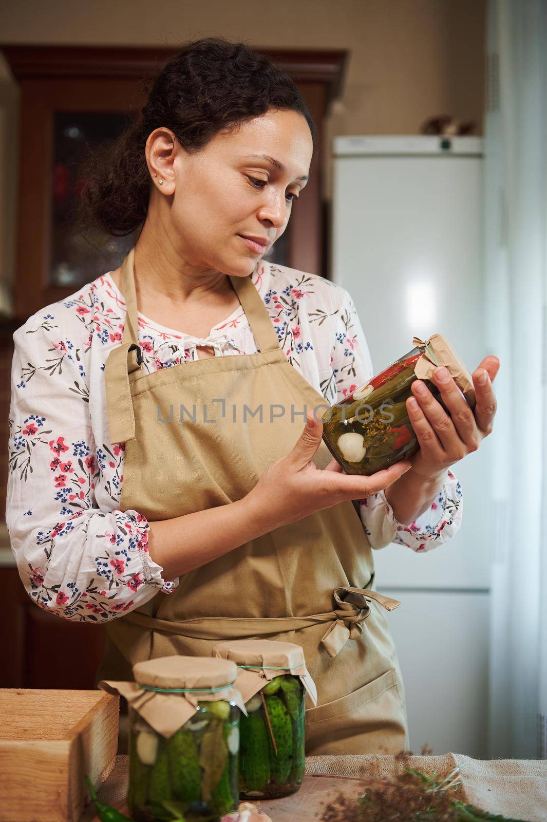 Delightful serene housewife looking at the jar with marinated chili peppers, she hold in her hands, standing by a kitchen table with cans of homemade pickled cucumbers and pickling ingredients