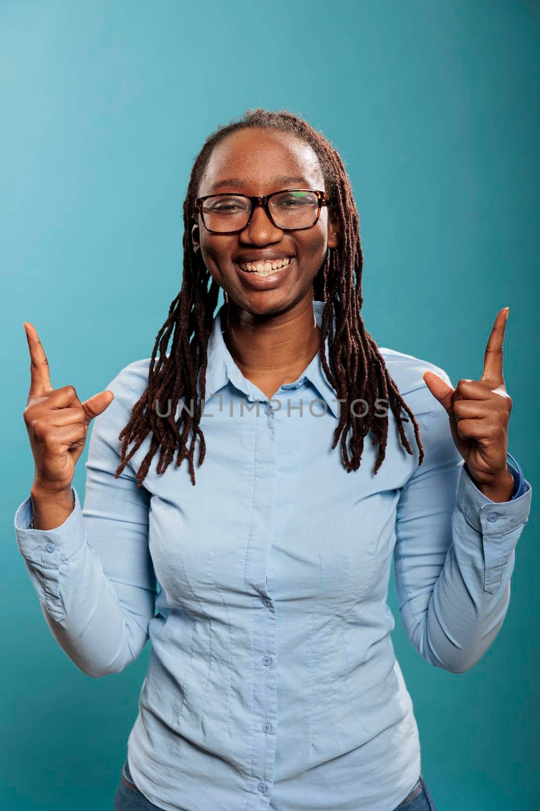 Excited happy beautiful woman smiling heartily while pointing fingers up on blue background. Confident and positive young adult person pointing hands up while looking at camera.