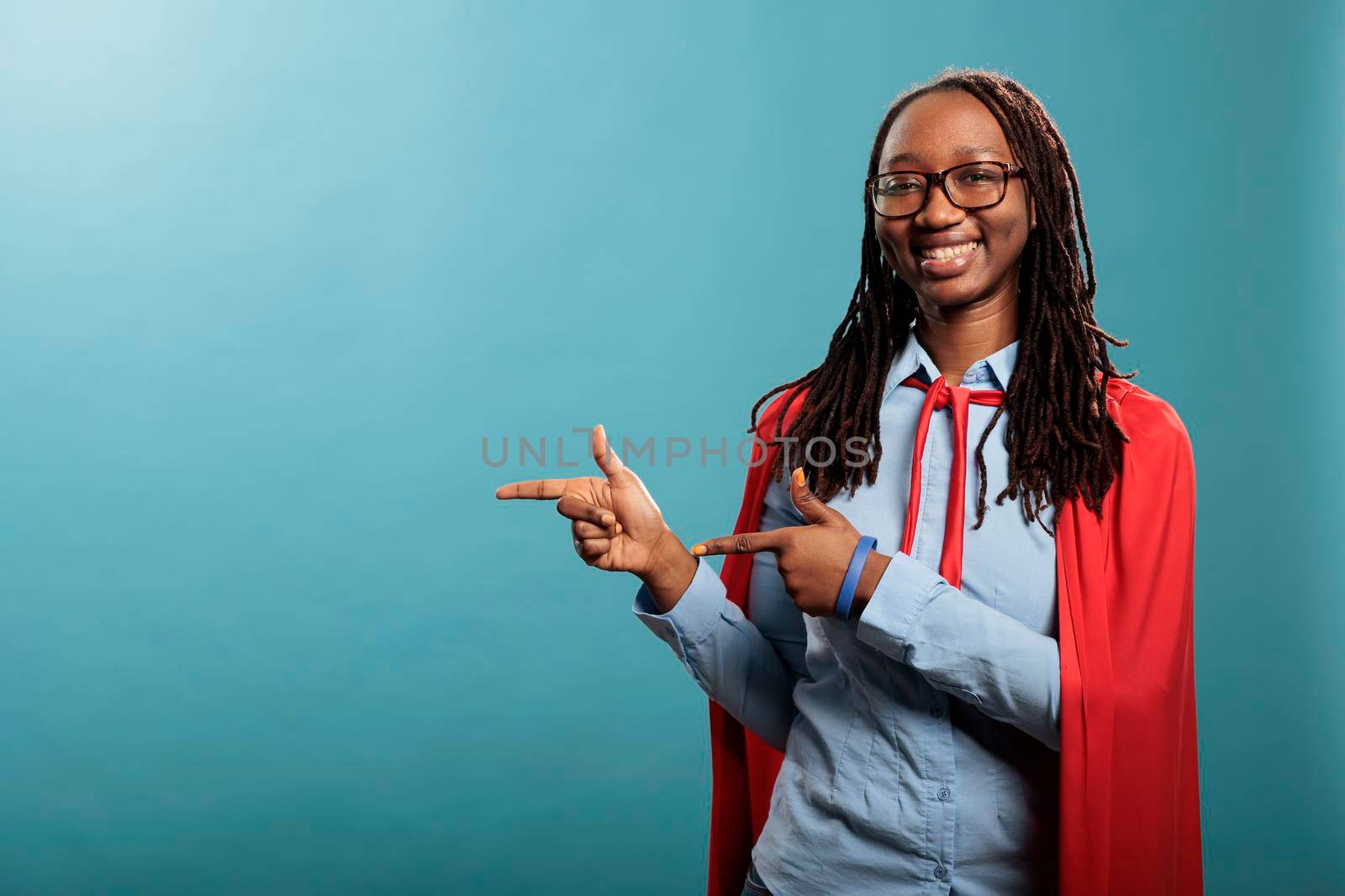 Brave and proud justice defender wearing mighty hero cape while pointing fingers to left on blue background. Positive and happy young adult justice defender person smiling heartily at camera.