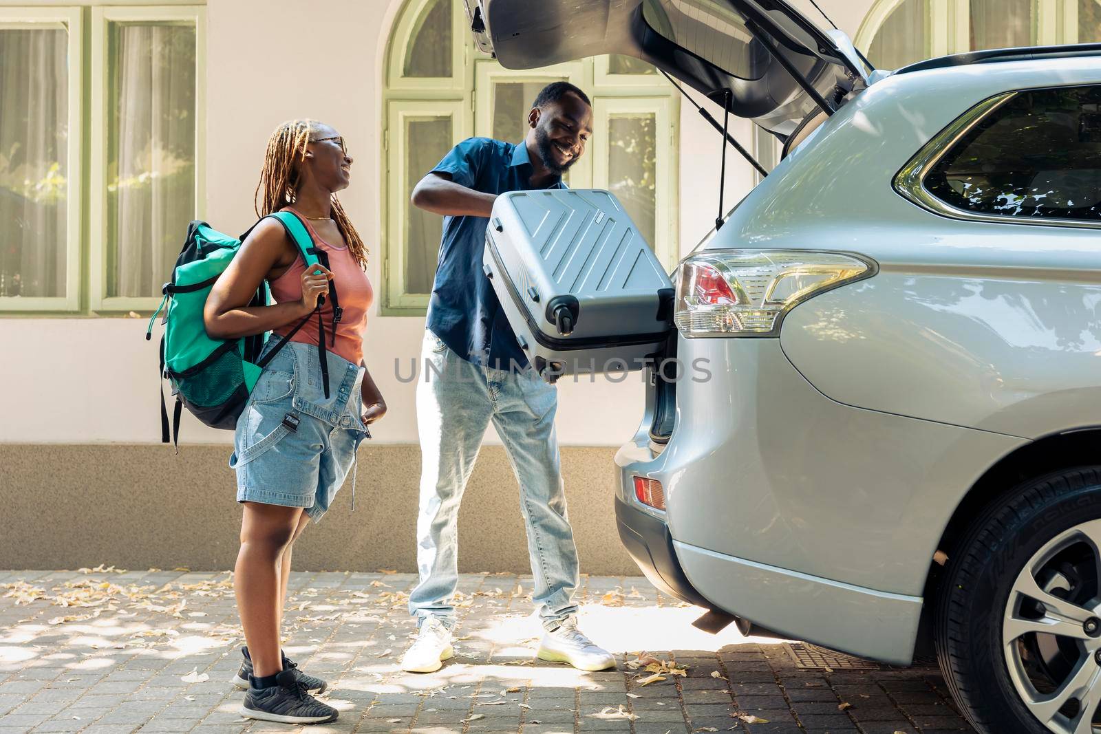 African american partners travelling by vehicle, putting luggage and baggage in trunk to leave on summer holiday. People in relationship going on vacation trip with trolley and travel bags.