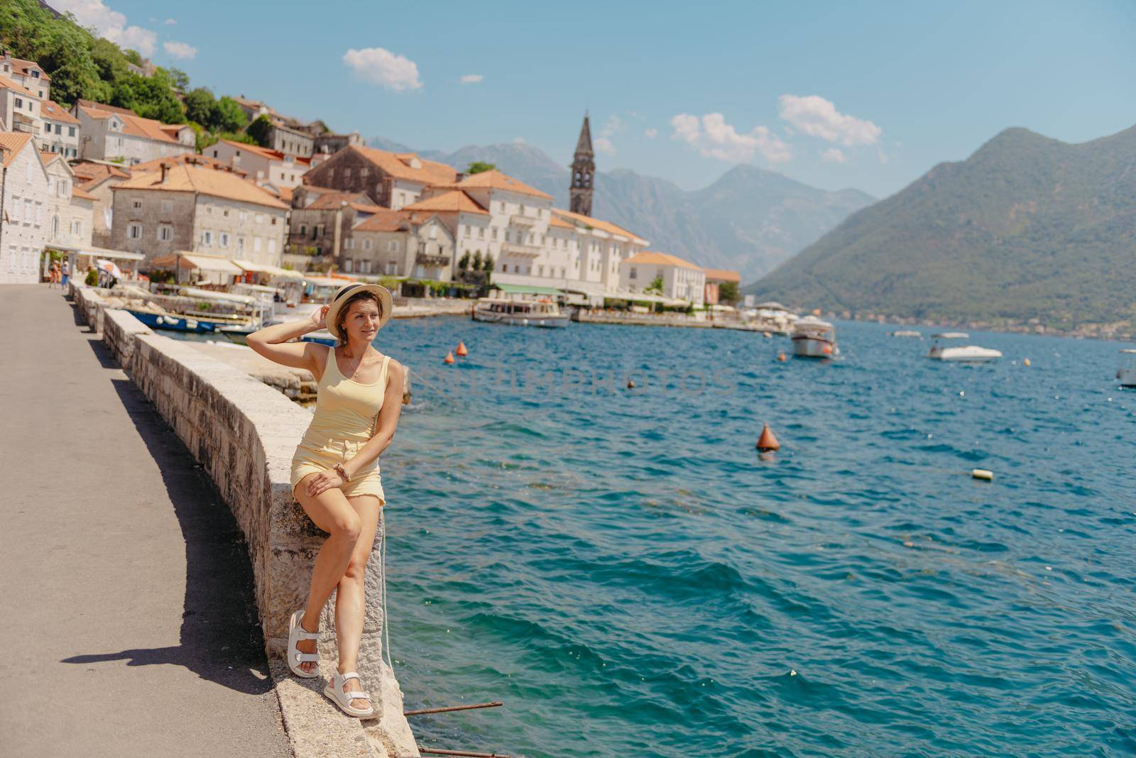 Summer photo shoot on the streets of Kotor, Montenegro. Beautiful girl in white dress and hat. smiling tourist girl with hat. Spectacular view of Montenegro with copy space. Ю fashion outdoor photo of beautiful sensual woman with blond hair in elegant dress and straw hat and bag, posing in Montenego's city Perast by Andrii_Ko