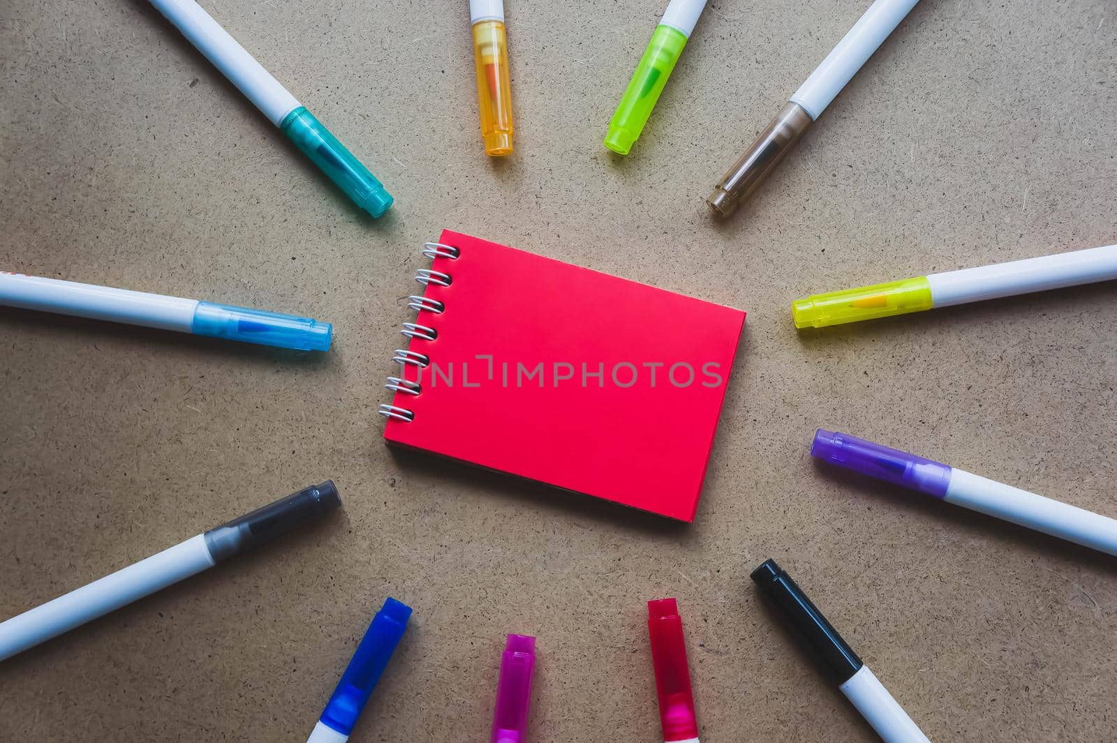 Notepad for notes with red sheets. Blank space for notes. Mocap. On warm ochre kraft background. Small spring-loaded notebook resting on table surrounded by markers. Children's creativity, school