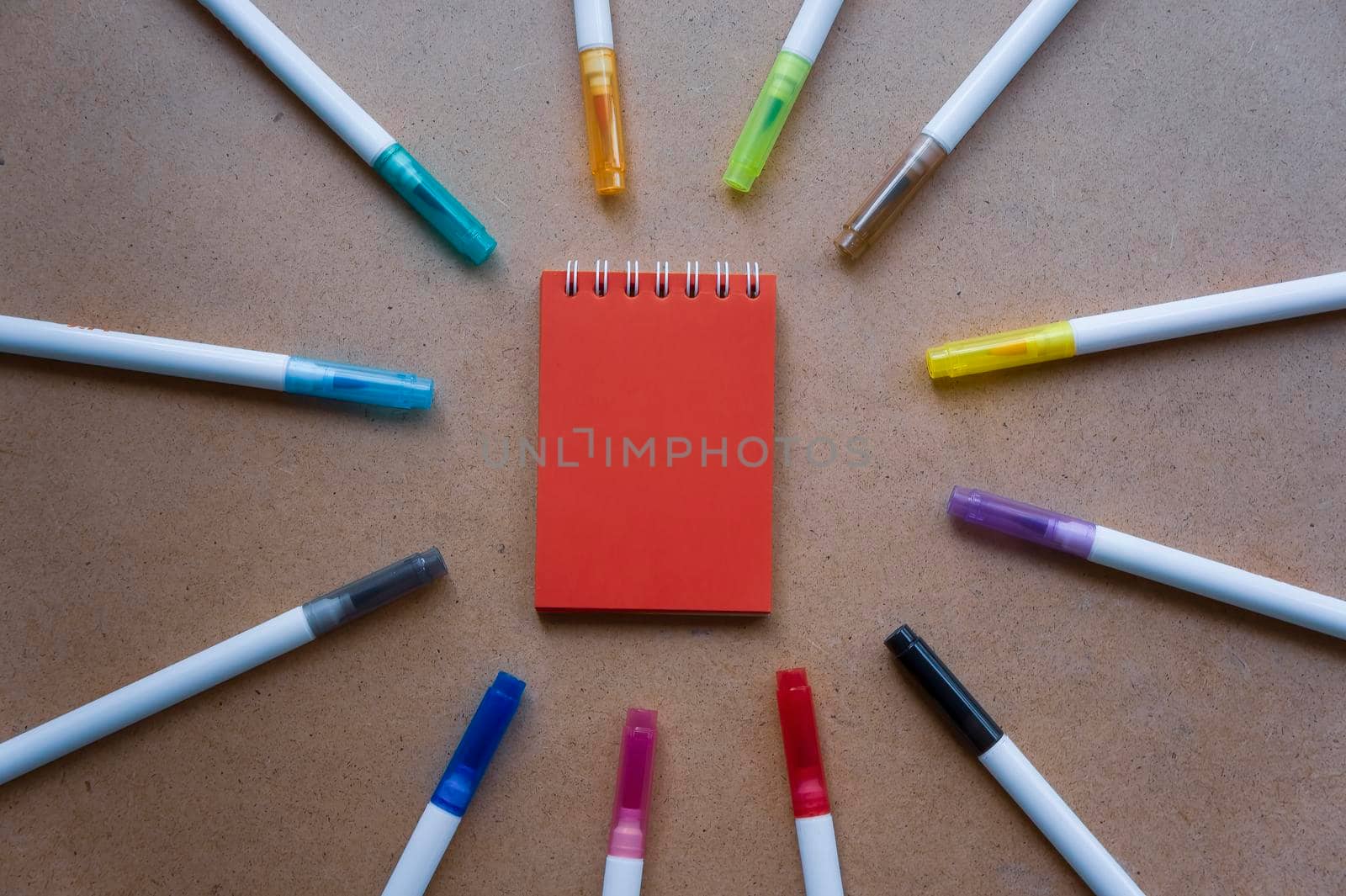 Notepad for notes with red sheets. Blank space for notes. Mocap. On warm ochre kraft background. Small spring-loaded notebook resting on table surrounded by markers. Children's creativity, school