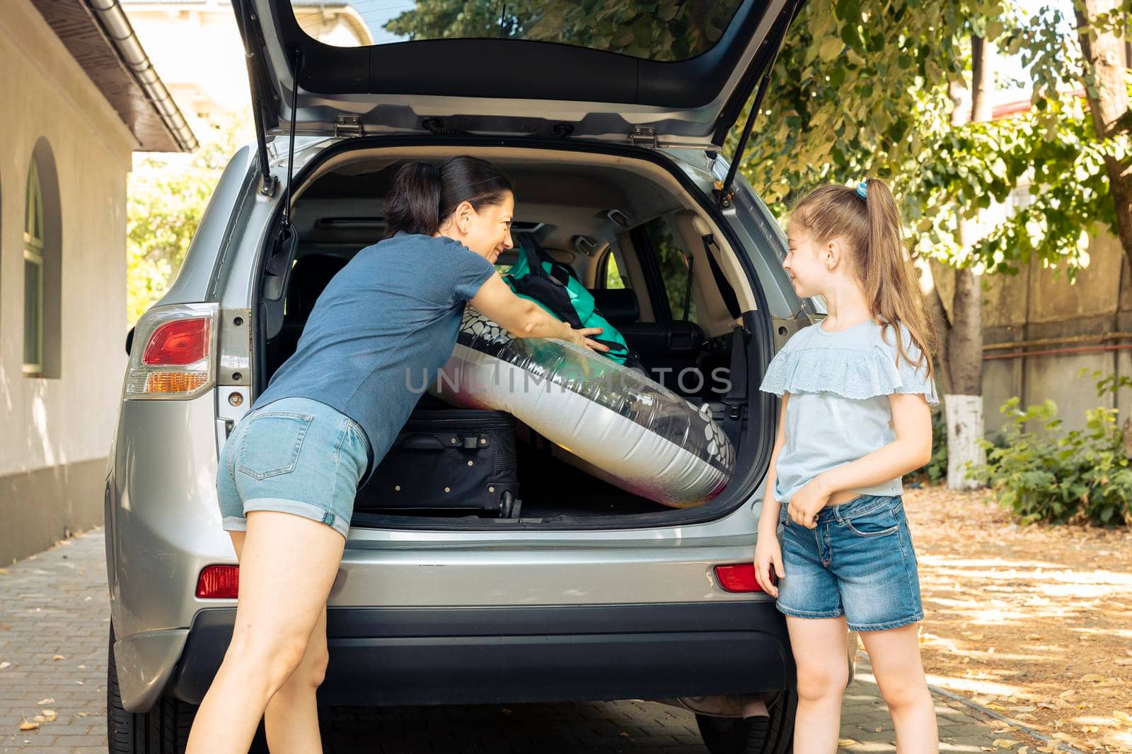 Mother and small kid loading baggage in automobile trunk to leave on holiday vacation at seaside. Travelling during summer with vehicle and luggage, suitcase, inflatable and trolley.