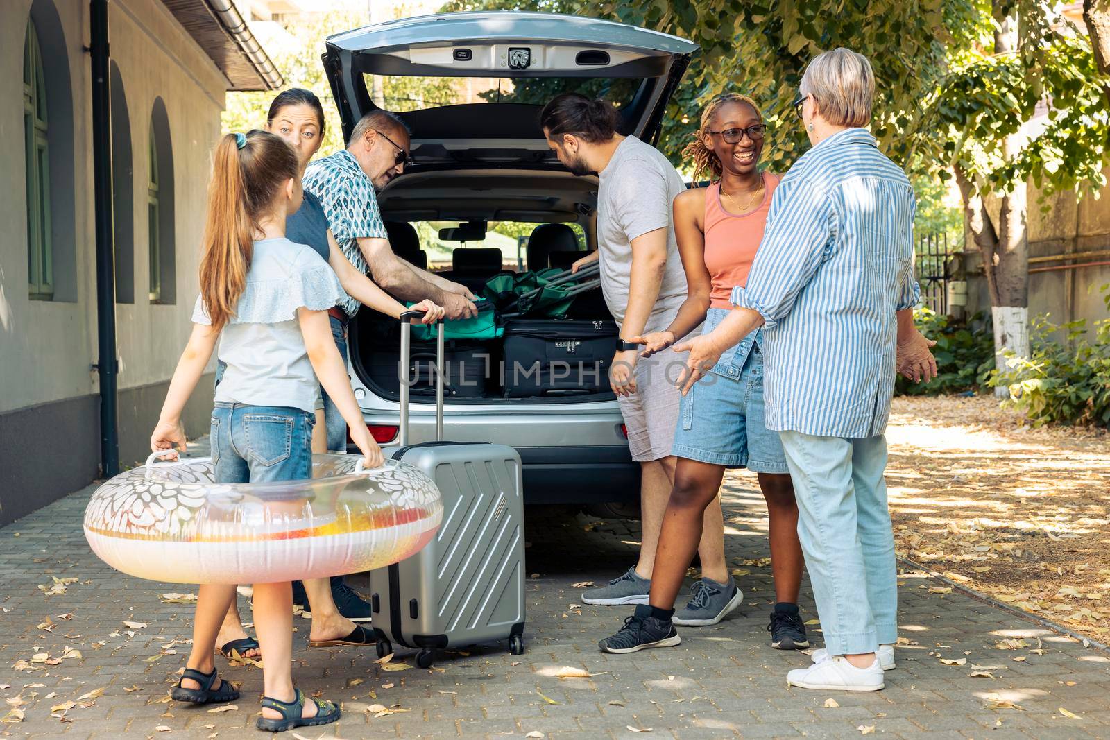 Multiethnic family and friends travelling on vacation, preparing to leave on summer holiday and loading baggage in car trunk. People sitting in driveway to go to seaside adventure trip.