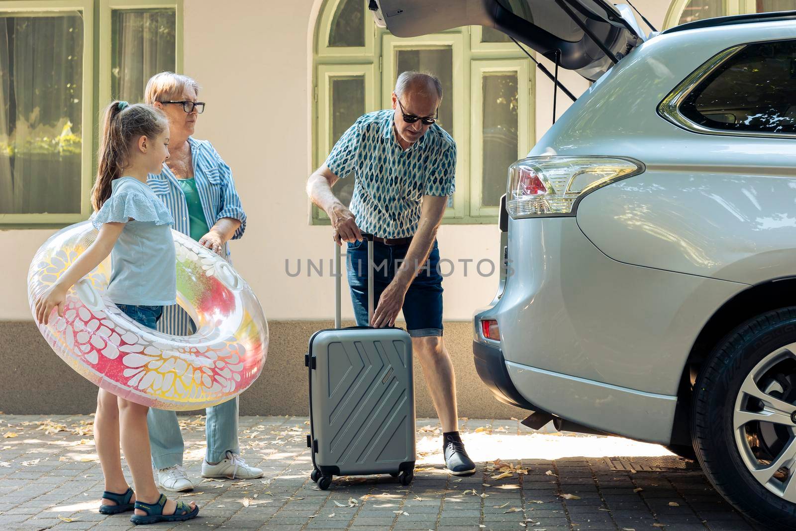 Elderly couple and niece loading baggage in trunk of automobile to leave on holiday road trip. Grandparents taking small child to seaside destination with luggage and inflatable during summer.
