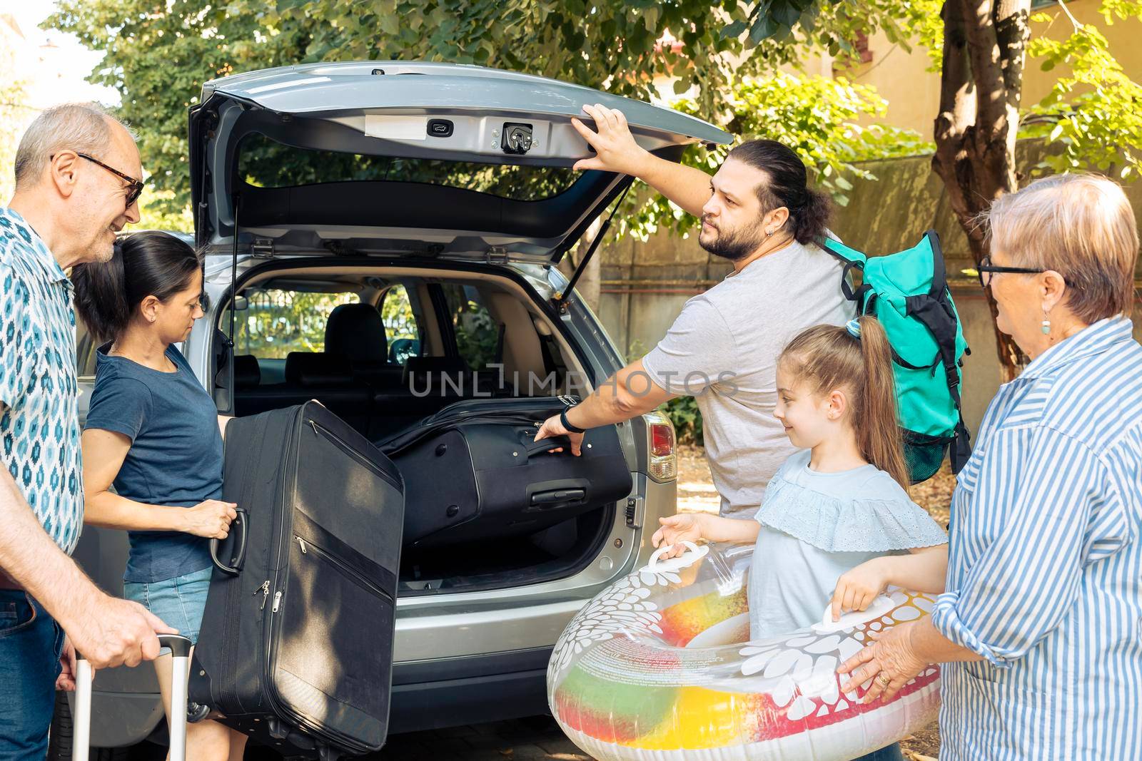 Big european family going on holiday vacation and loading baggage in automobile trunk. Child travelling with parents and grandparents at seaside during summer, putting luggage in vehicle.