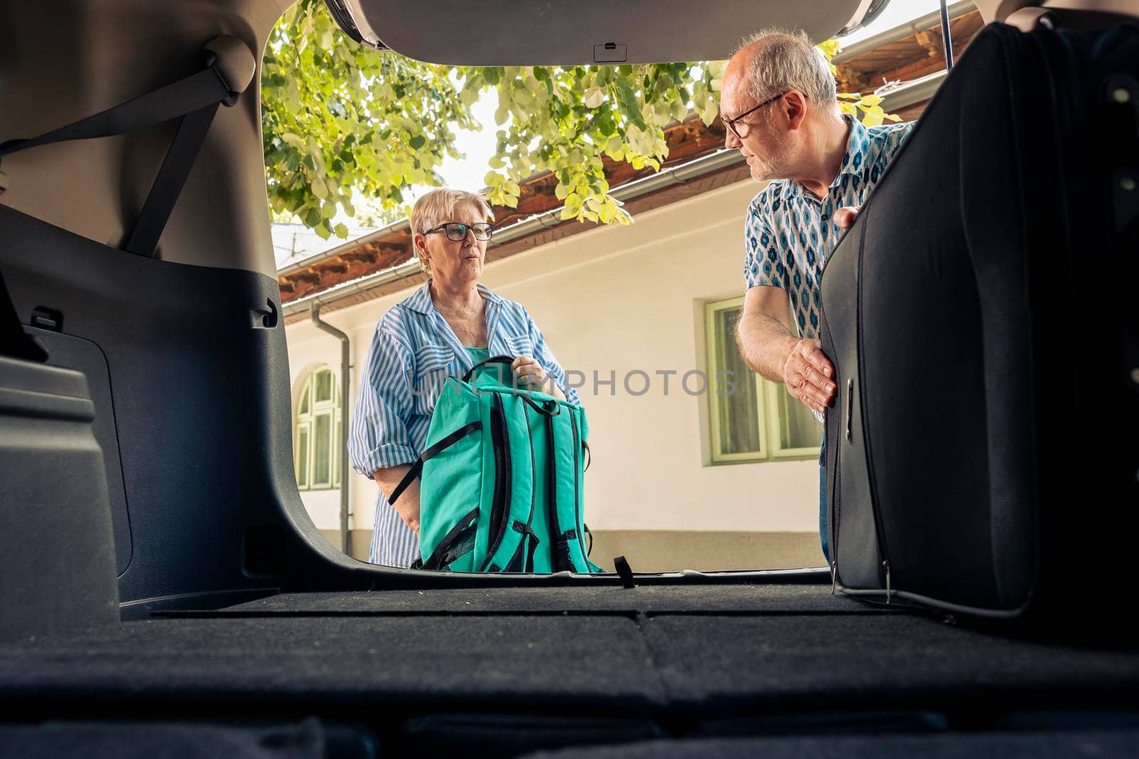 Senior people loading baggage in car trunk, preparing to leave on summer holiday with vehicle. Couple travelling on vacation road trip in retirement age, going on adventure destination.