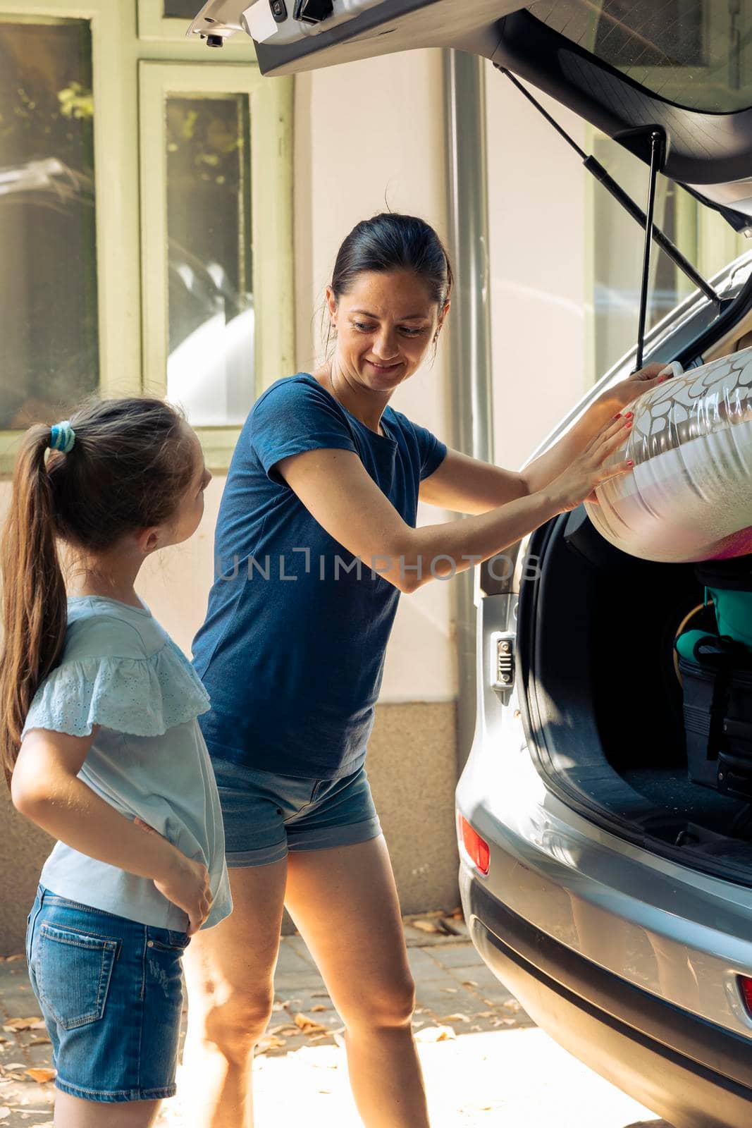 Mother and little girl going to seaside with vehicle, travelling together on road trip adventure. Family leaving on summer holiday vacation with inflatable, travel bags and trolley in trunk.