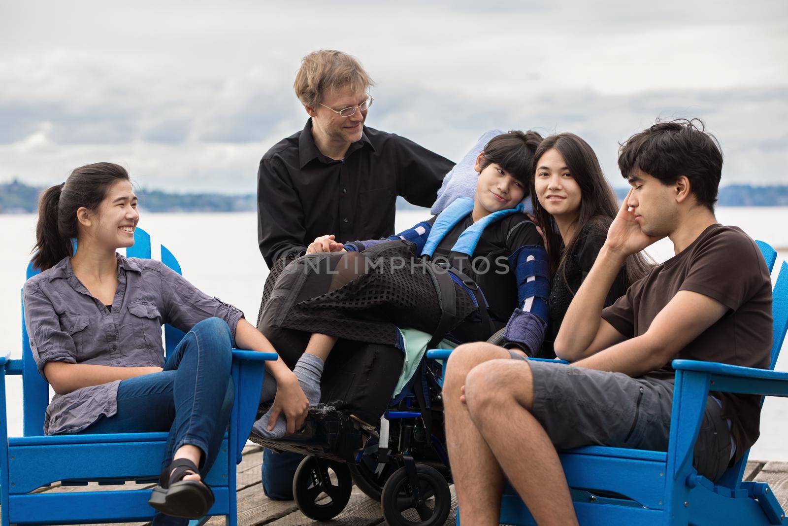 Family of multiethnicity with child who has special needs outdoors together on summer day. Child is sitting in wheelchair.