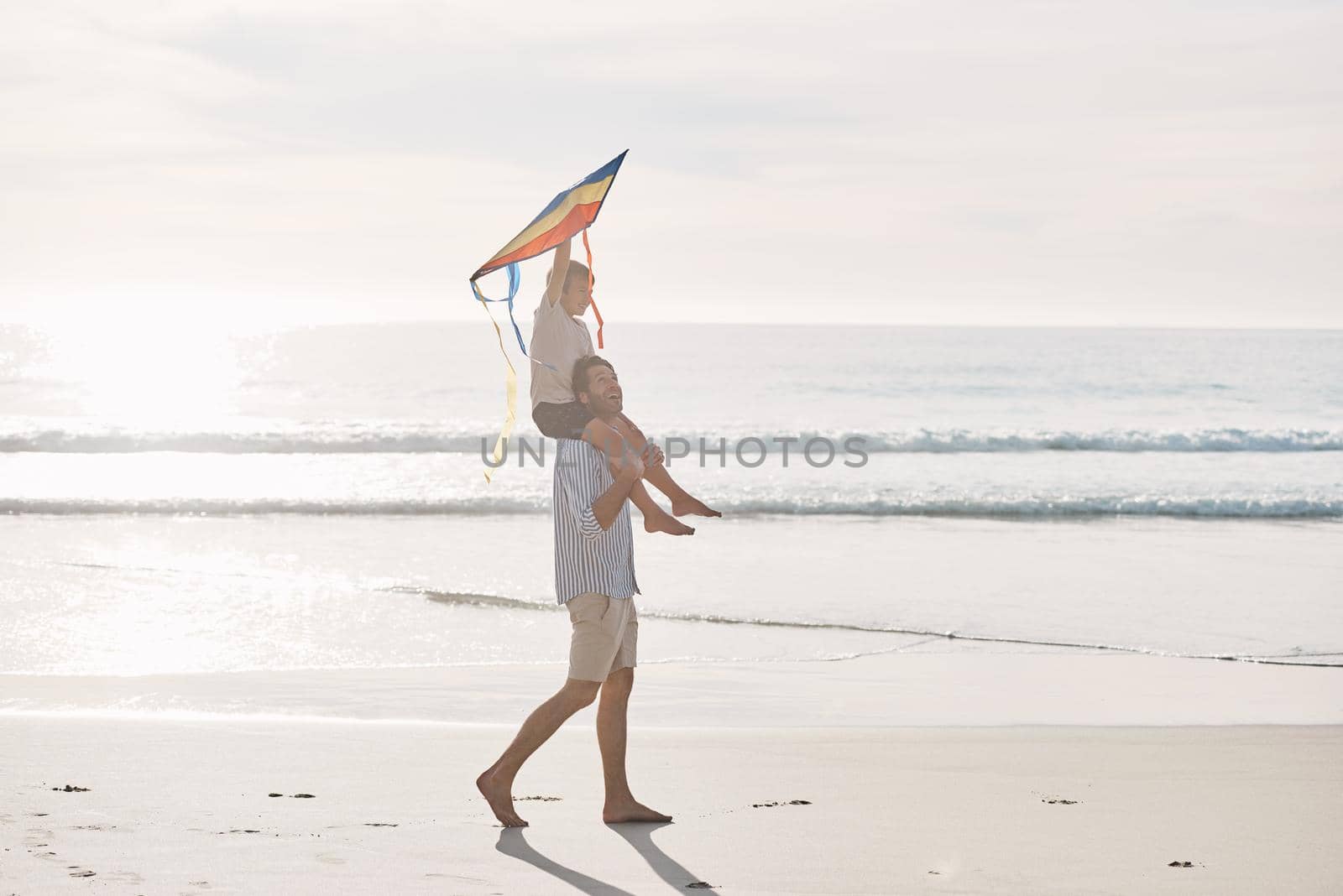 Hold it high, son. Full length shot of a young boy being carried on his fathers shoulders and holding a kite on the beach. by YuriArcurs
