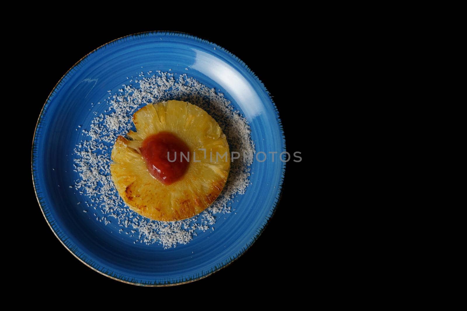 close-up of a grilled natural pineapple with grated coconut on a blue plate on a black background