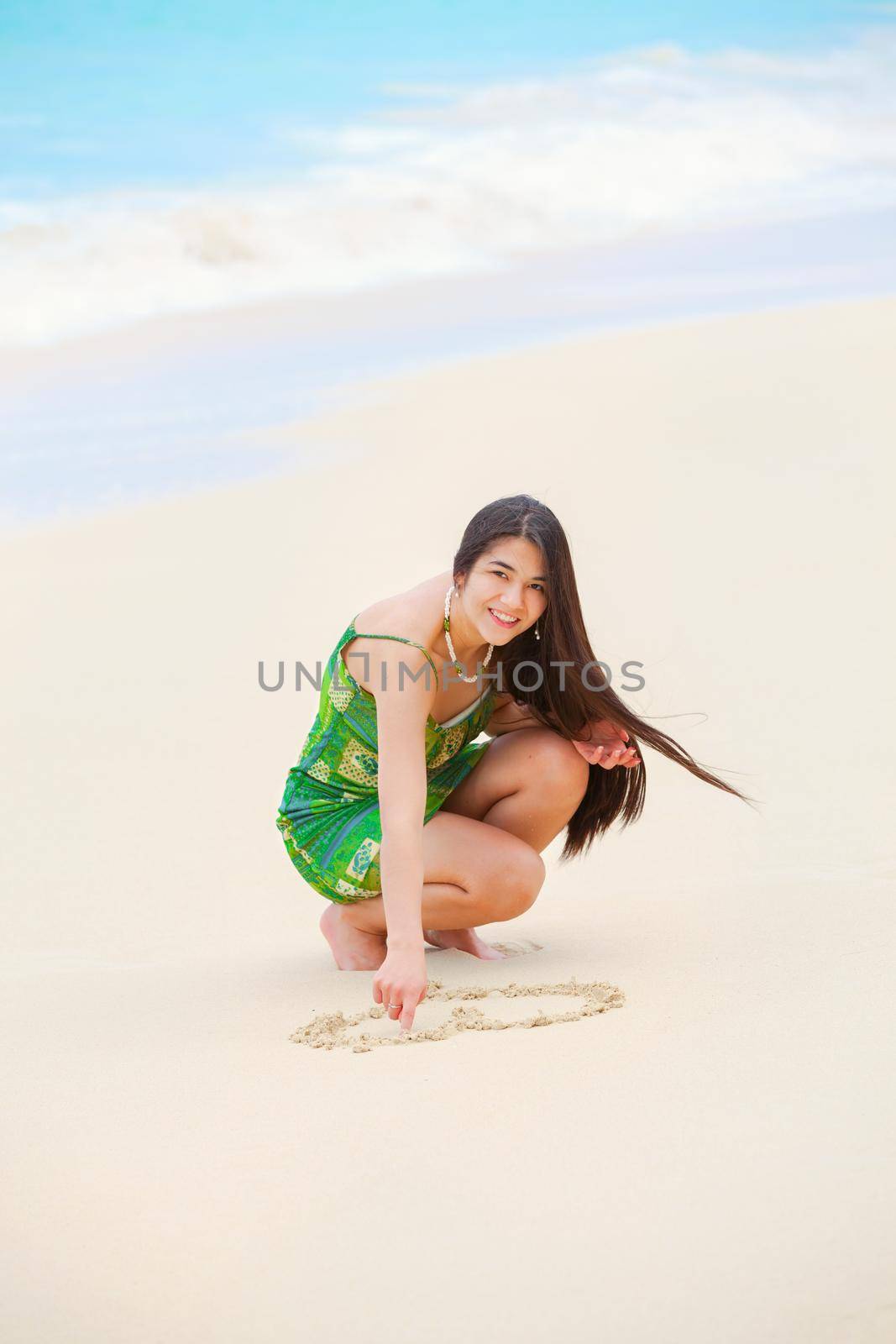 Beautiful teen girl crouching down, drawing heart in sand on tropical beach