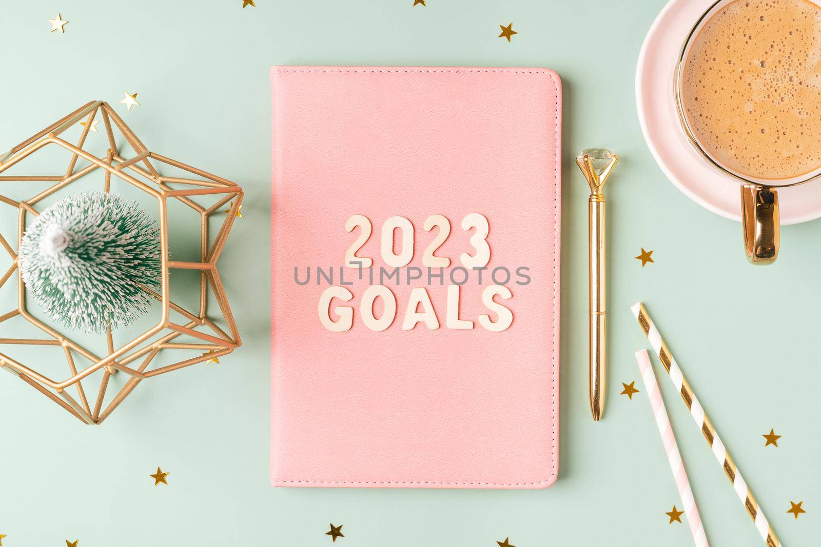 Christmas pink notepad with 2023 goals letters text. Flat lay on green mint table background with planner, cup of coffee, candle, Christmas decoration, notebook and stationery. Top view Desktop.