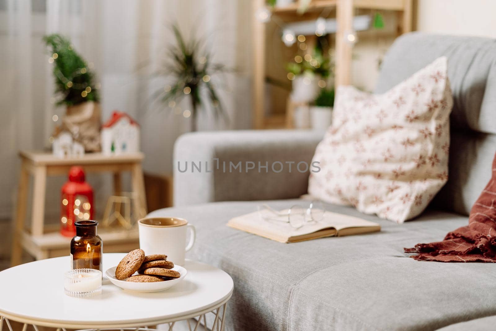 Stylish Christmas room interior with Christmas light and decor. Winters holiday with cup of hot coca or coffee, cookies, book and candles. Cozy home.
