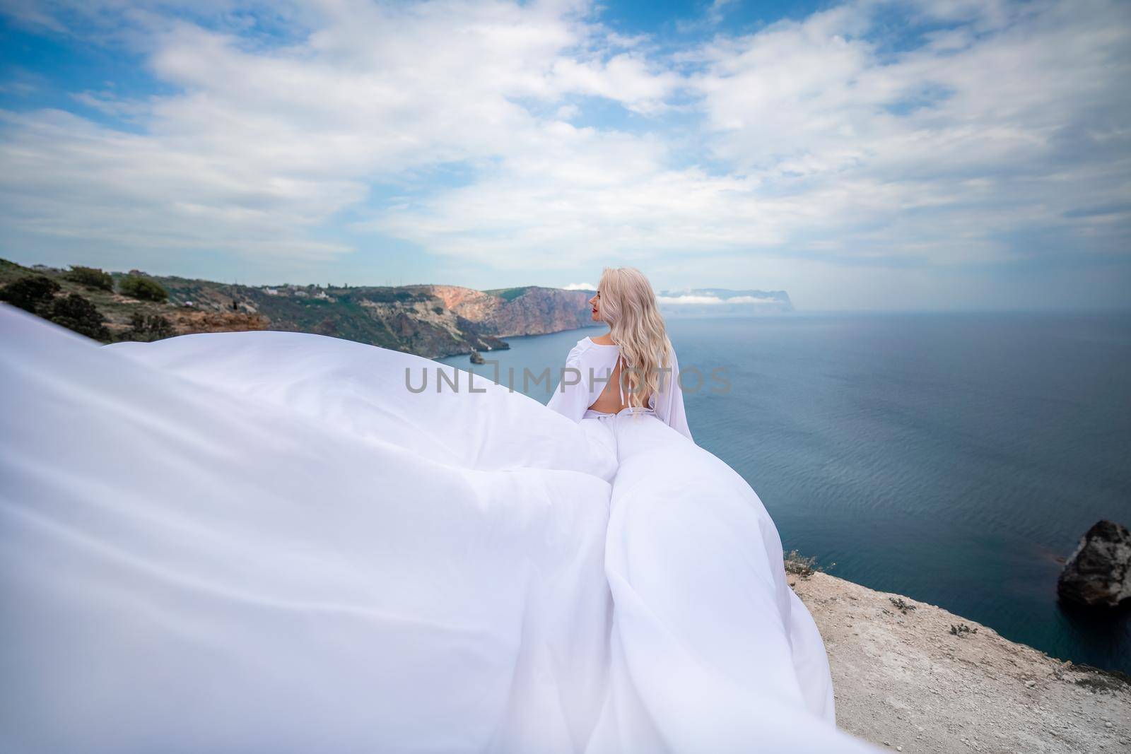 Blonde with long hair on a sunny seashore in a white flowing dress, rear view, silk fabric waving in the wind. Against the backdrop of the blue sky and mountains on the seashore