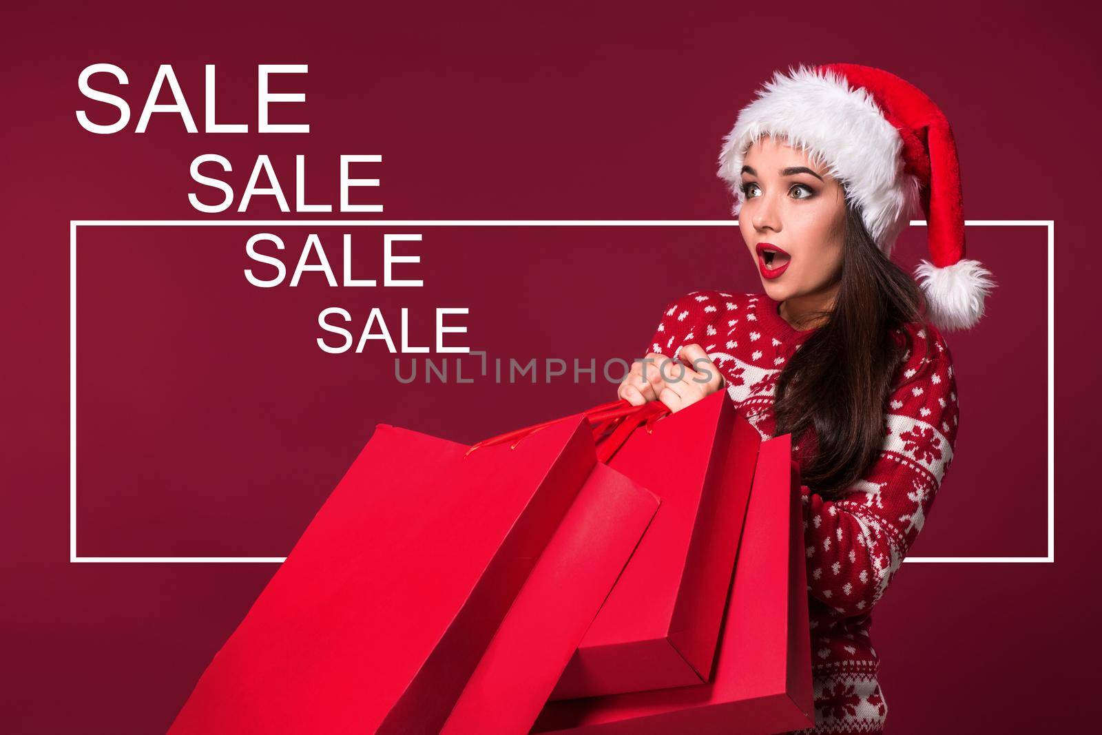 Christmas woman. Beauty model girl in santa hat on red background. Funny laughing surprised woman portrait. Open mouth. True emotions. Red lips and beautiful holiday makeup. Shopping and sale. Shopaholic girl. Black Friday, holiday celebration.