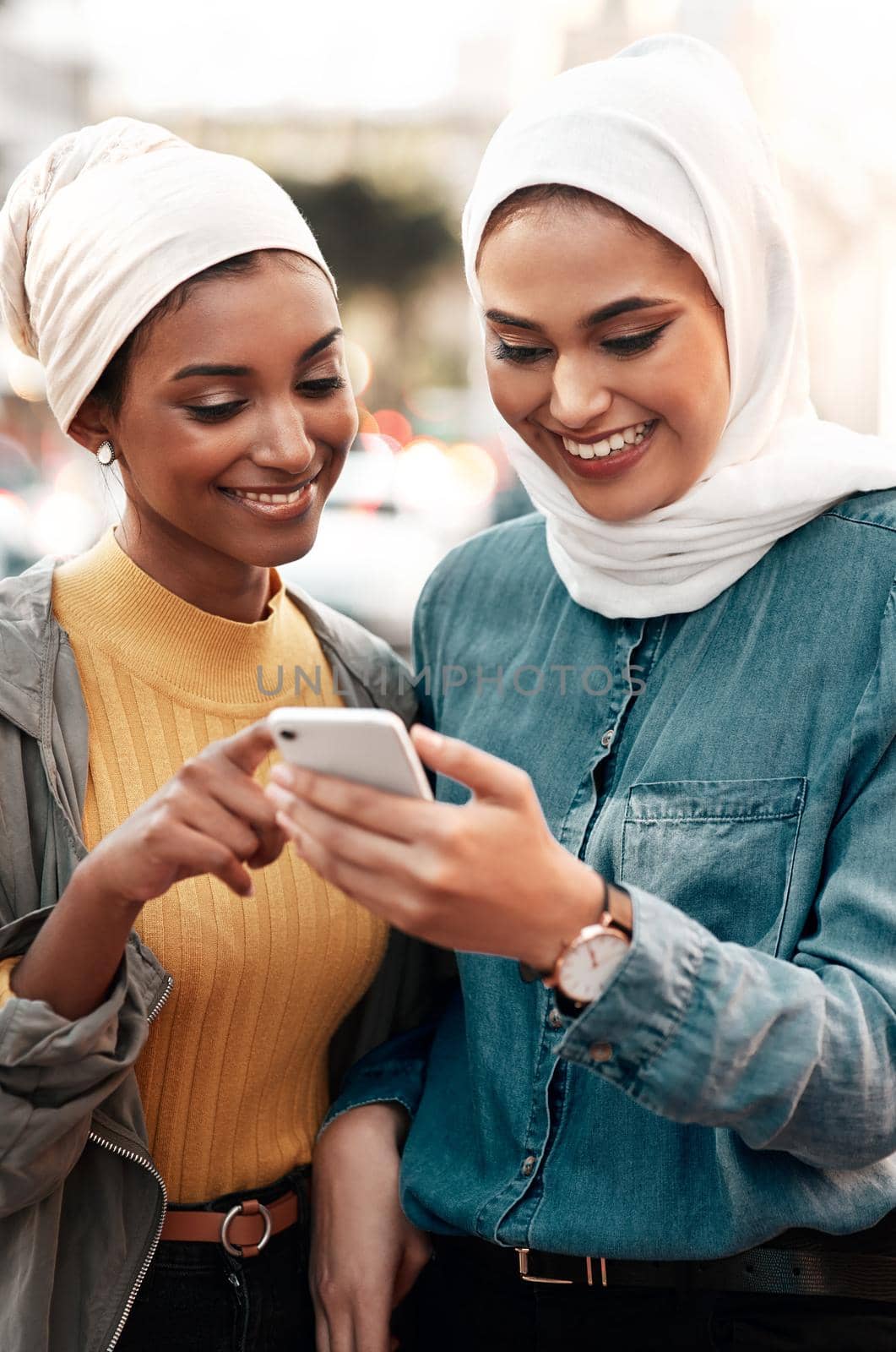 This is where we should go. two attractive young women wearing headscarves and standing together while using a cellphone in the city. by YuriArcurs