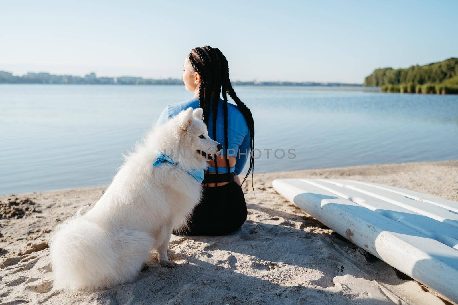 Woman with Locs Sitting on Beach of City Lake with Her Best Friend, Dog Breed Japanese Spitz, and Preparing for Paddleboarding