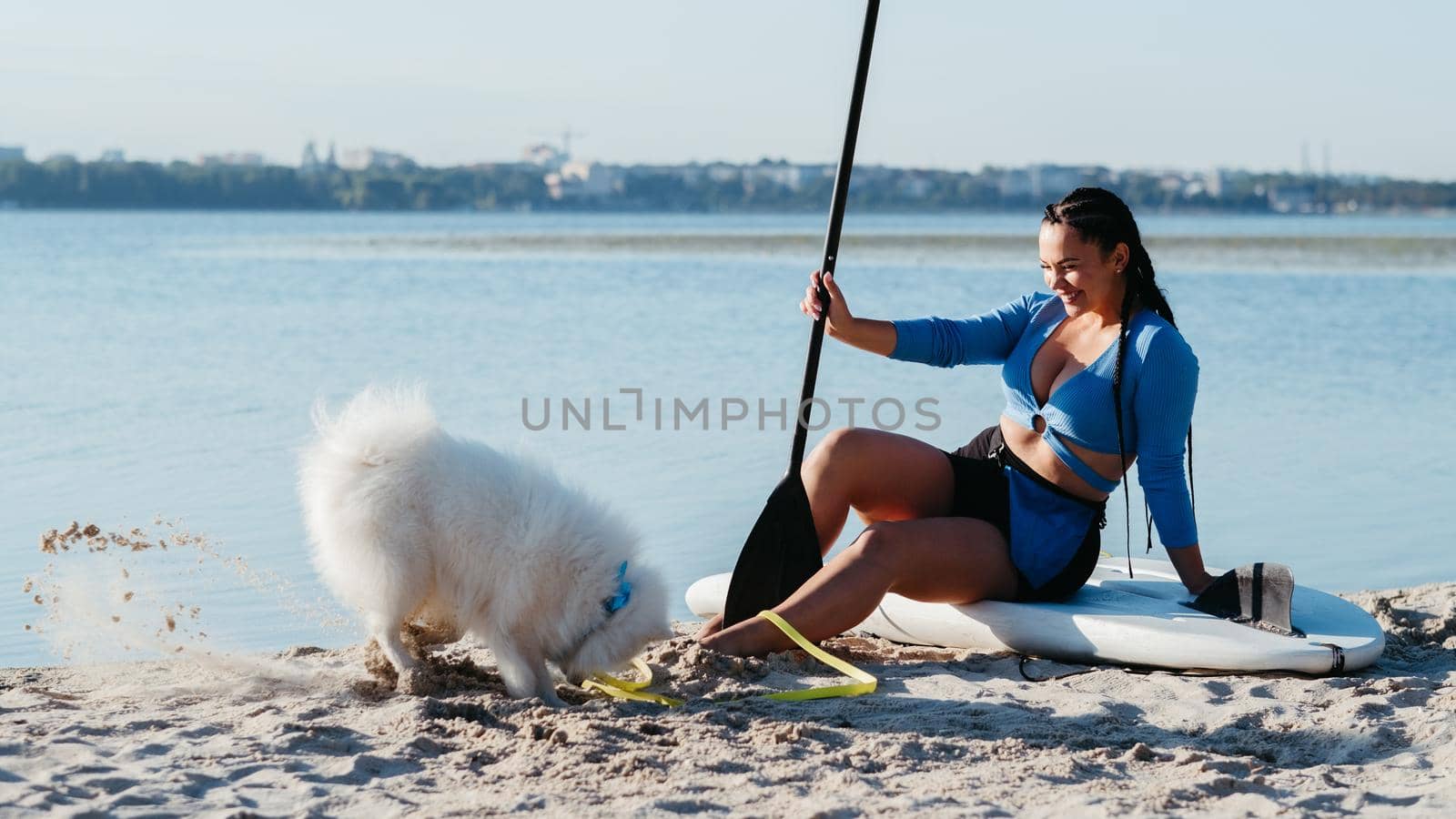 Young Woman Sitting on Sup Board and Laughing at Her Japanese Spitz Digging a Hole on the Beach of City Lake, Paddleboarding with Dog by Romvy