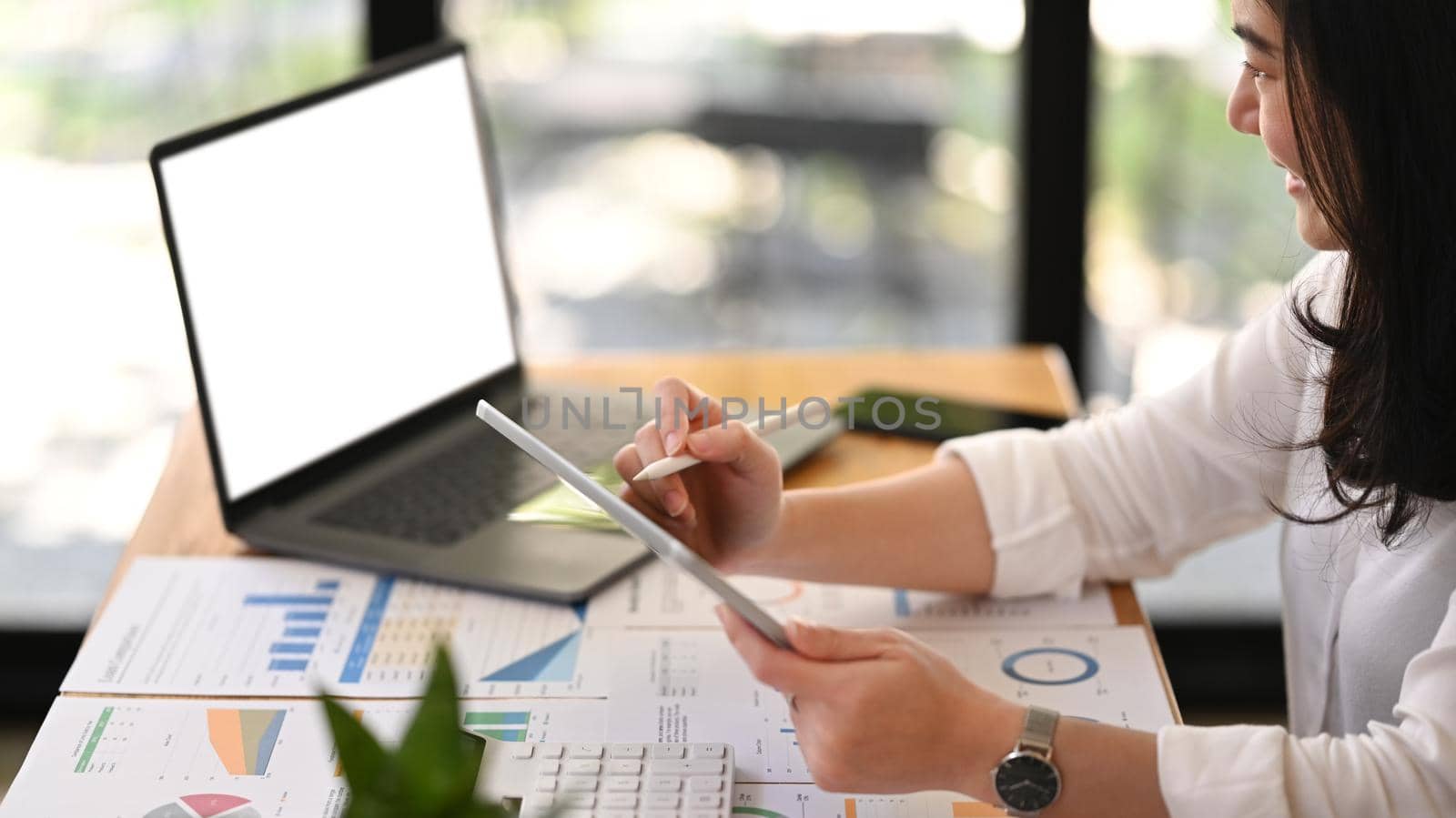 Side view of smiling young employee working with modern devices and document at office desk.