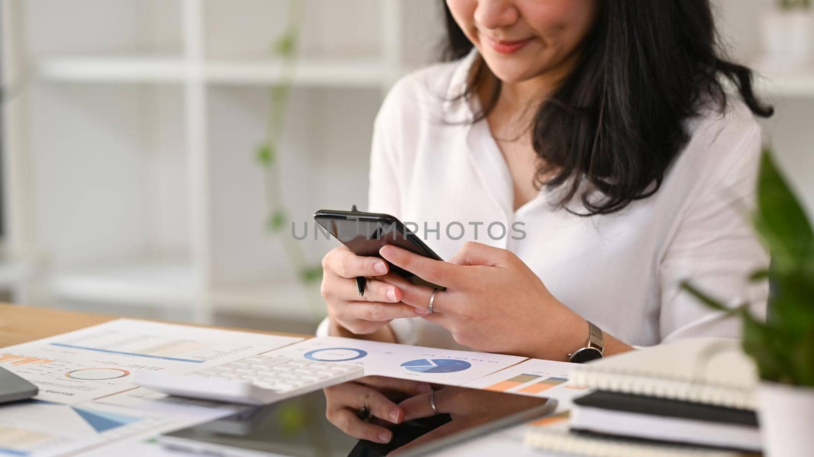 Smiling female office worker communicating in social media or chatting online on mobile phone.