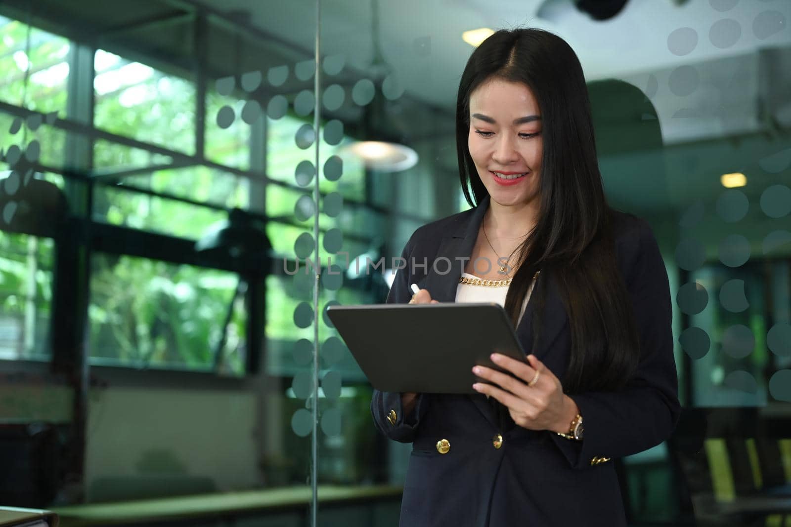 Smiling female team leader standing in corporate office and using digital tablet.