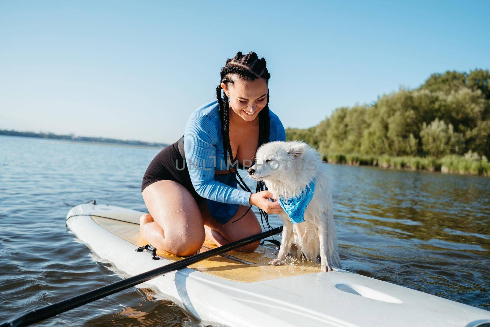 Cheerful Woman with Locs Helping Her Dog Japanese Spitz Get Out of the Water, While Sitting on Sup Board on the Lake