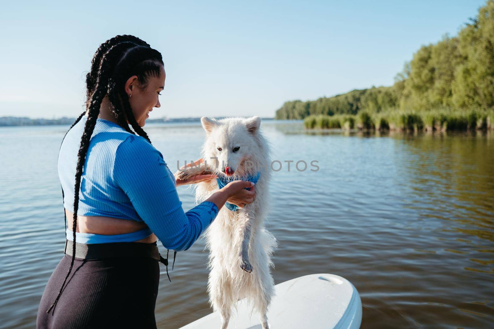 Young Woman with Locs Holding Her Pet Japanese Spitz on the Lake, Dog Standing on Sup Board on His Back Paws