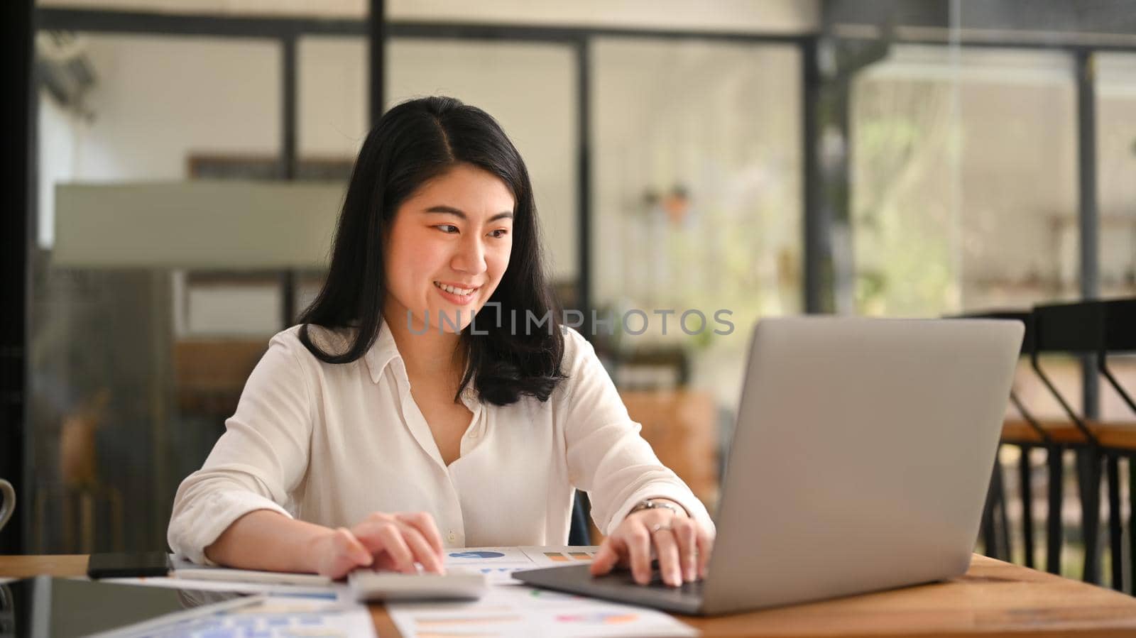 Charming female economist using laptop computer and calculator at corporate office.