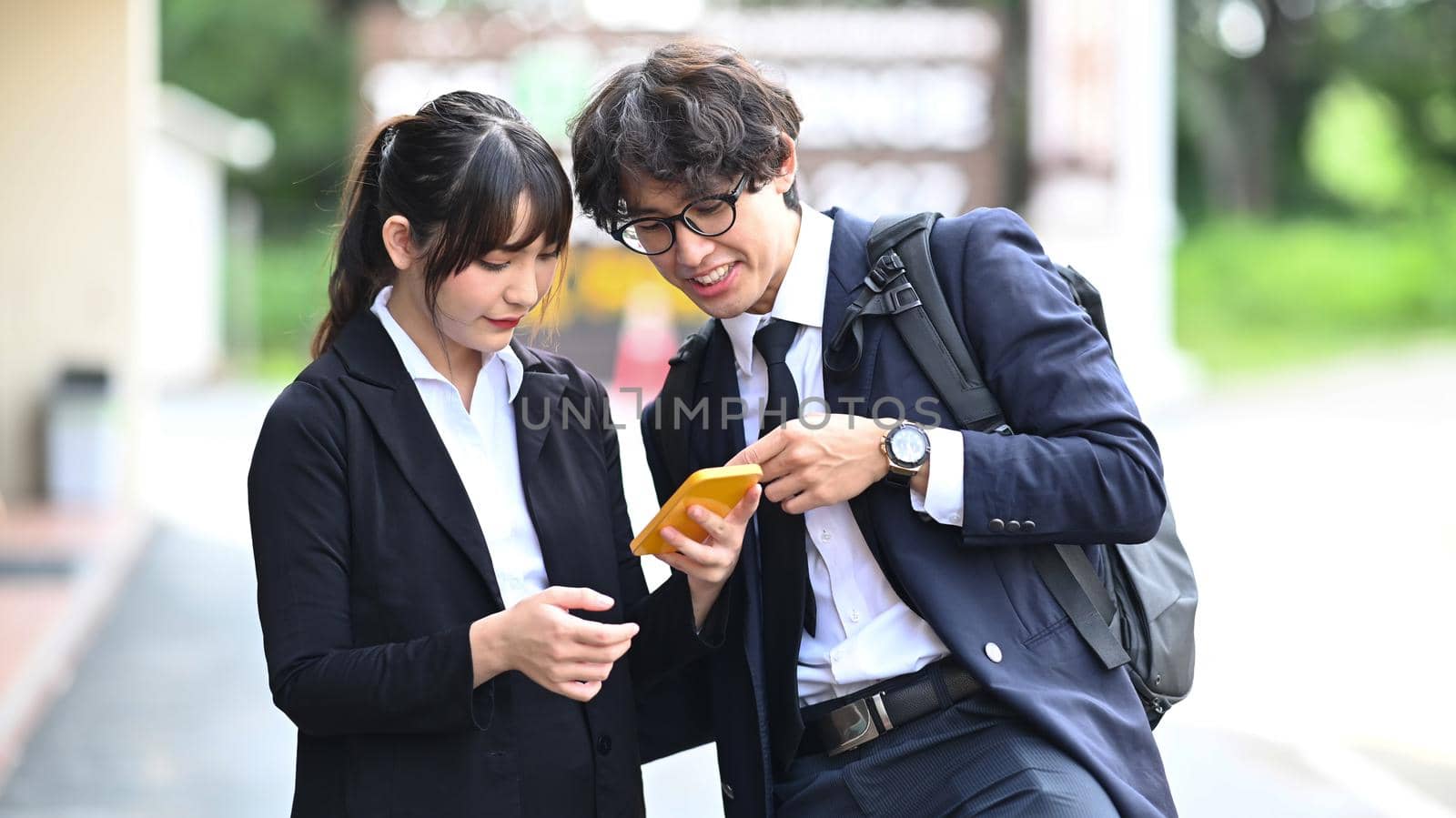 Two business colleagues using smart phone while standing outdoors at the city streets. by prathanchorruangsak