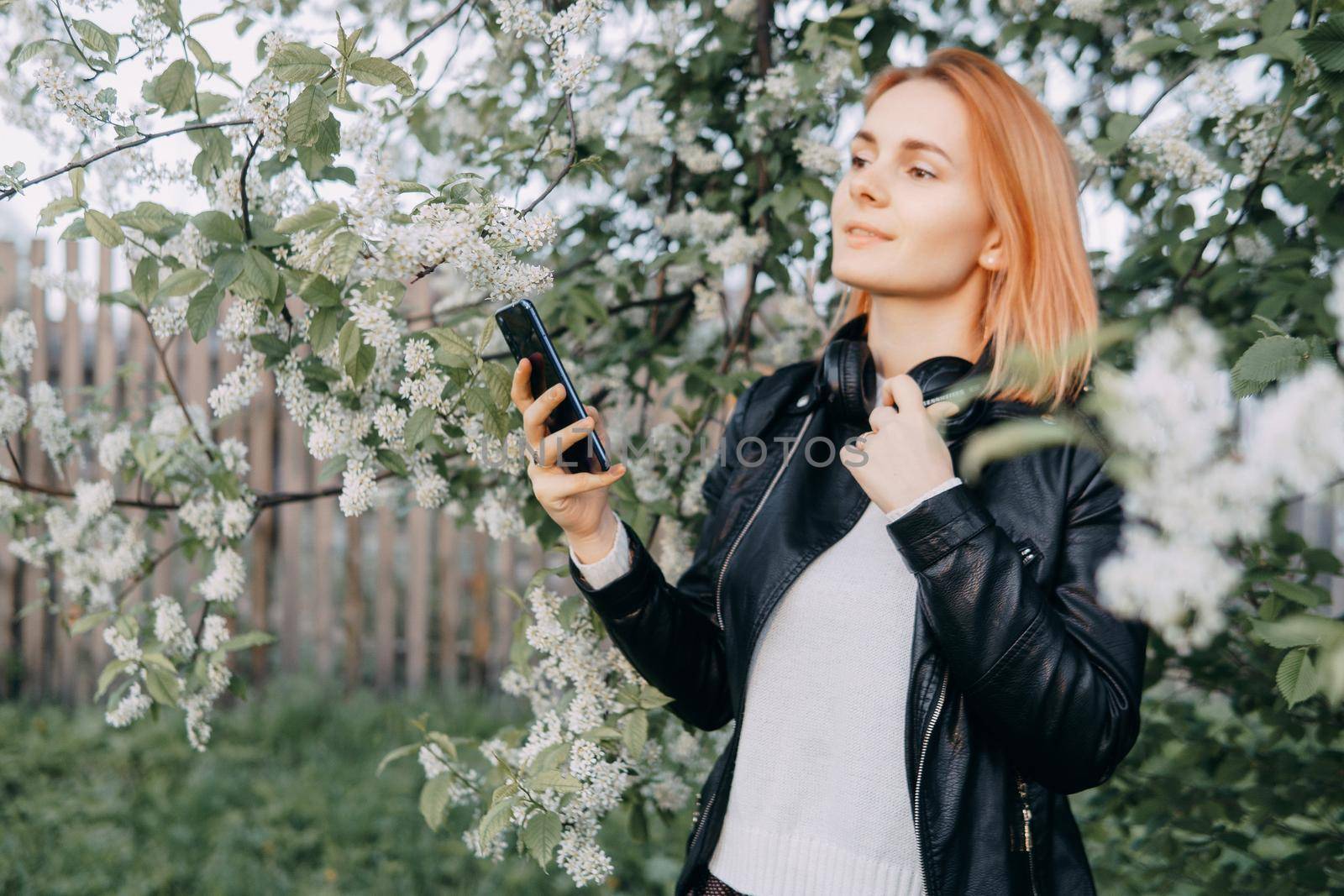 A red-haired teenage girl with headphones around her neck takes a selfie against the background of a flowering cherry tree. The time of spring flowering of cherry orchards.