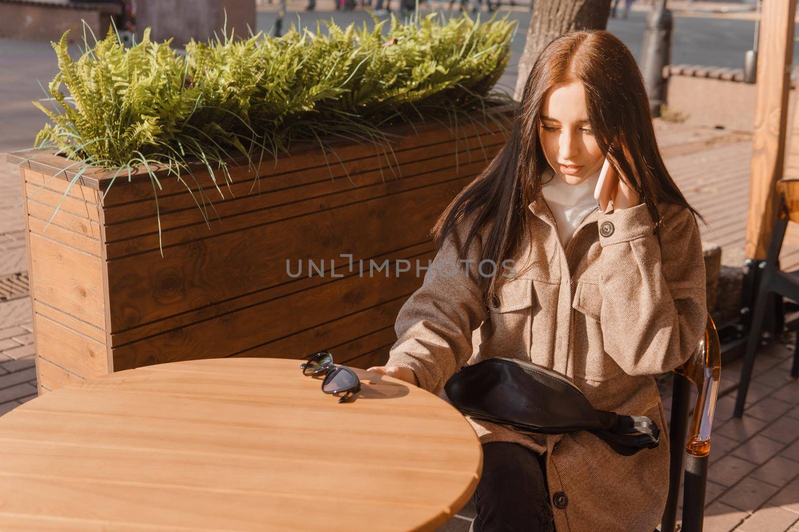 A stylish brunette woman in sunglasses is sitting at a table in an outdoor cafe with coffee and a phone. The girl is talking on the phone. Autumn walk.
