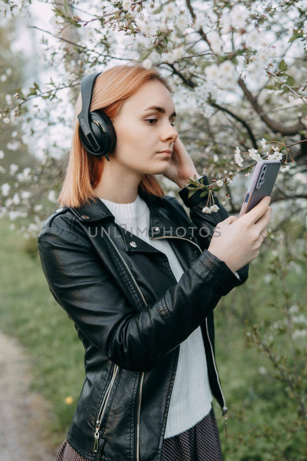 A red-haired teenage girl listens to music in headphones, configured via the phone. The time of spring flowering of cherry orchards.