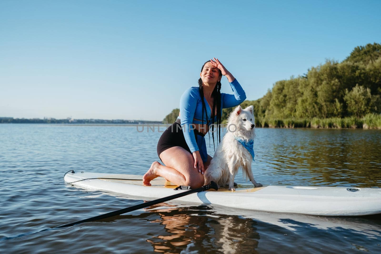 Happy Young Woman Enjoying Life on the Lake at Early Morning Sitting on Sup Board with Her Dog Japanese Spitz