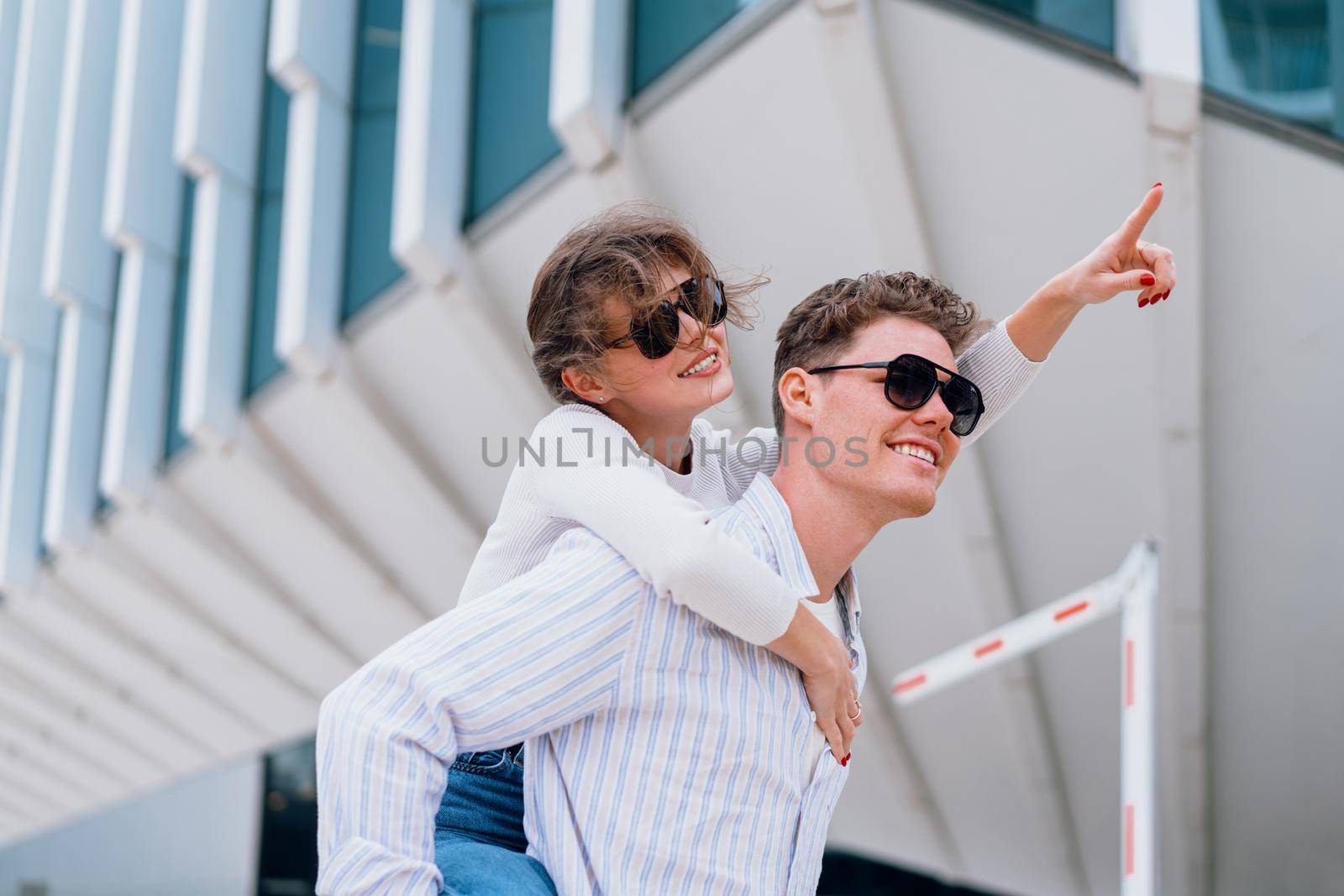 Carefree young urban couple in sunglasses doing piggyback at city street outdoor with modern commercial building on background. Man carrying young woman on his back, woman show by finger away