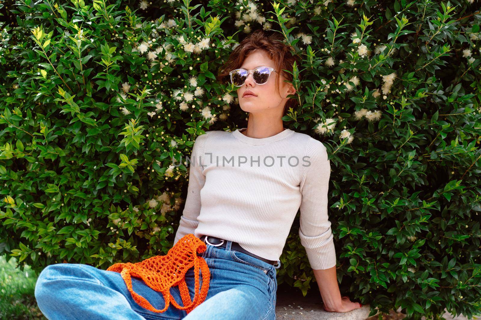 Young woman in light summer clothes with a reusable mesh eco bag and sunglasses, sitting bench at summer park before shopping. Sustainable lifestyle. Eco friendly concept.