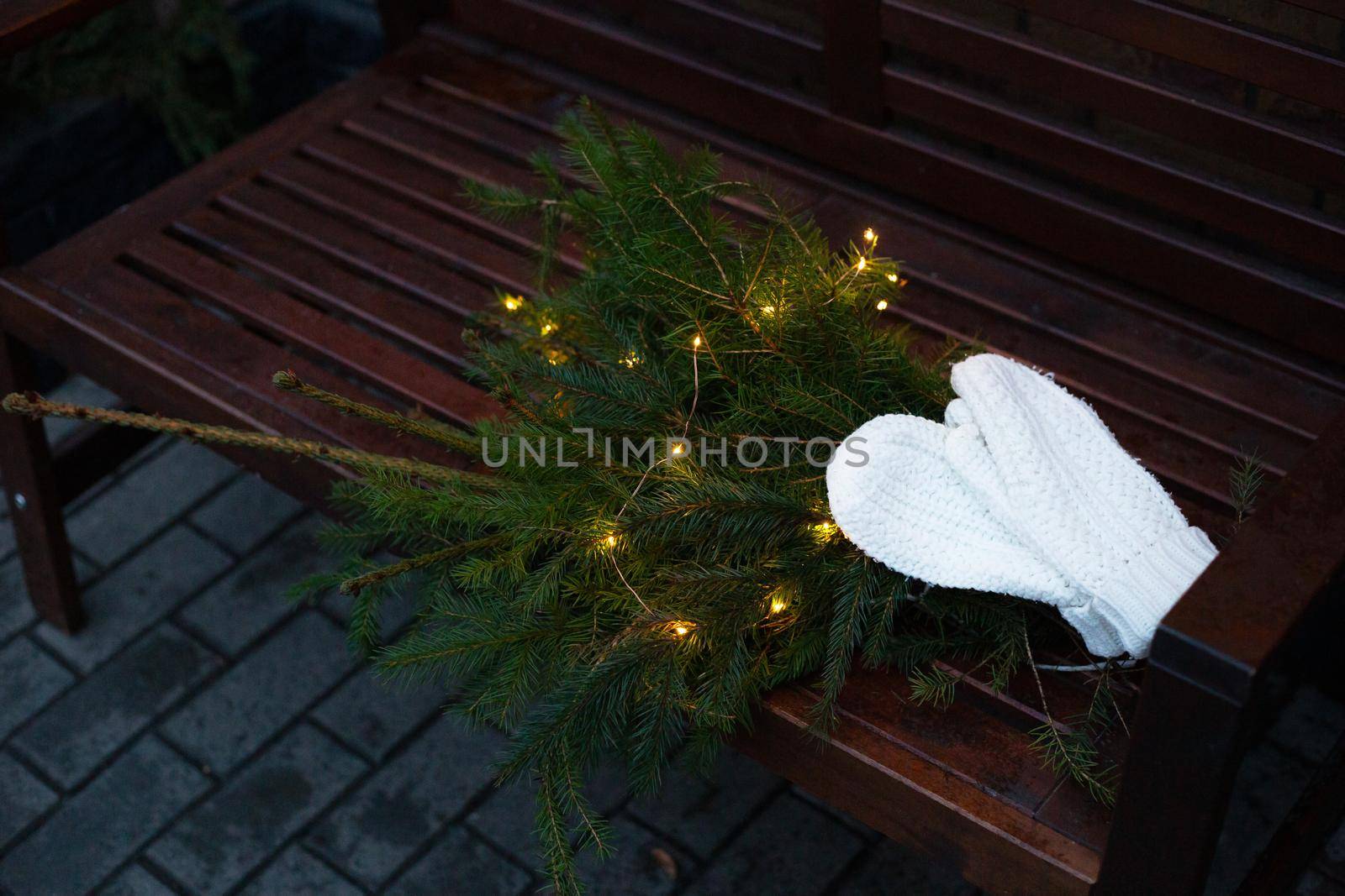 Branches of a coniferous tree decorated with a garland lie on a bench along with knitted white mittens