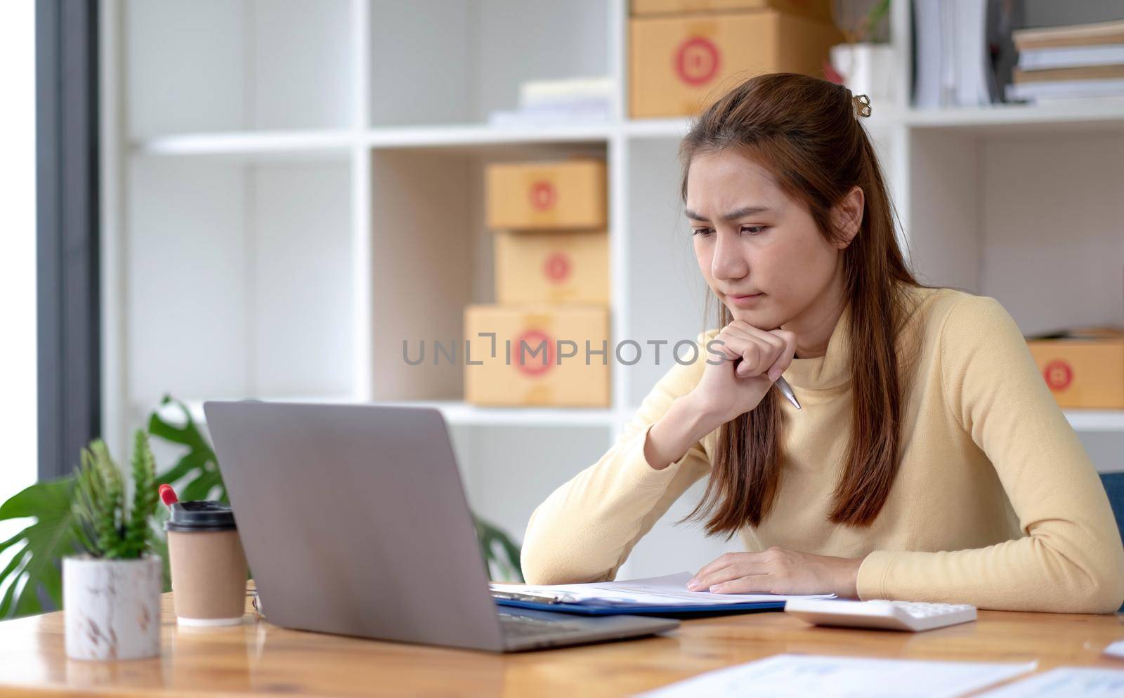 A portrait of young serious Asian woman working with laptop in the office full of packages and boxes stacking up, busy looking table, for SME, delivery, start up business and home office concept..