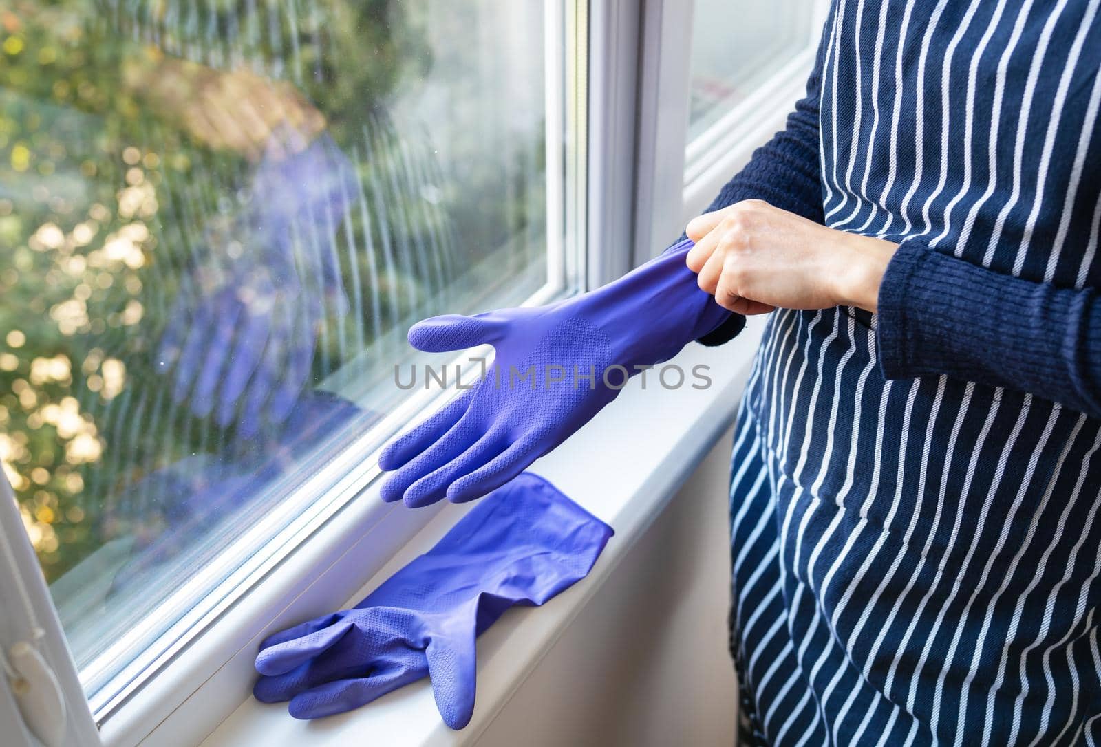 A young girl in a striped apron puts purple gloves on her hands. To prepare for washing windows in an apartment or house. Cleaning and cleaning concept. by sfinks