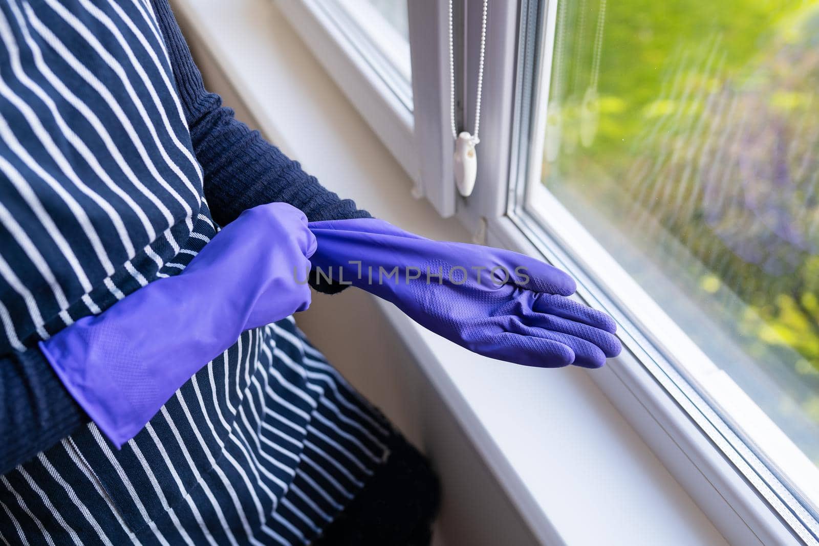 A young girl in a striped apron puts purple gloves on her hands. To prepare for washing windows in an apartment or house. Cleaning and cleaning concept
