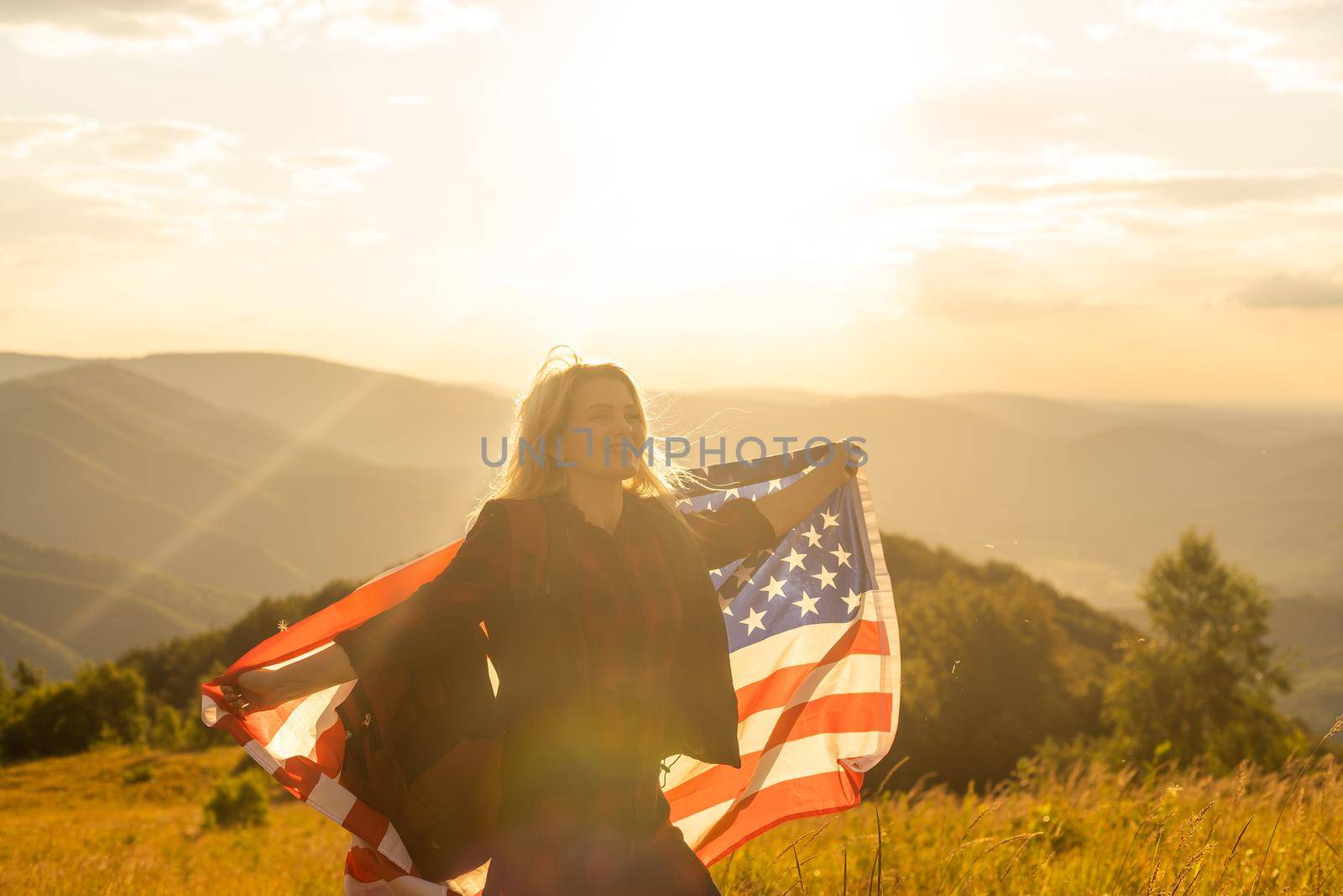 Young happy american woman with long hair holding waving on wind USA national flag on her sholders relaxing outdoors enjoying warm summer day by Andelov13
