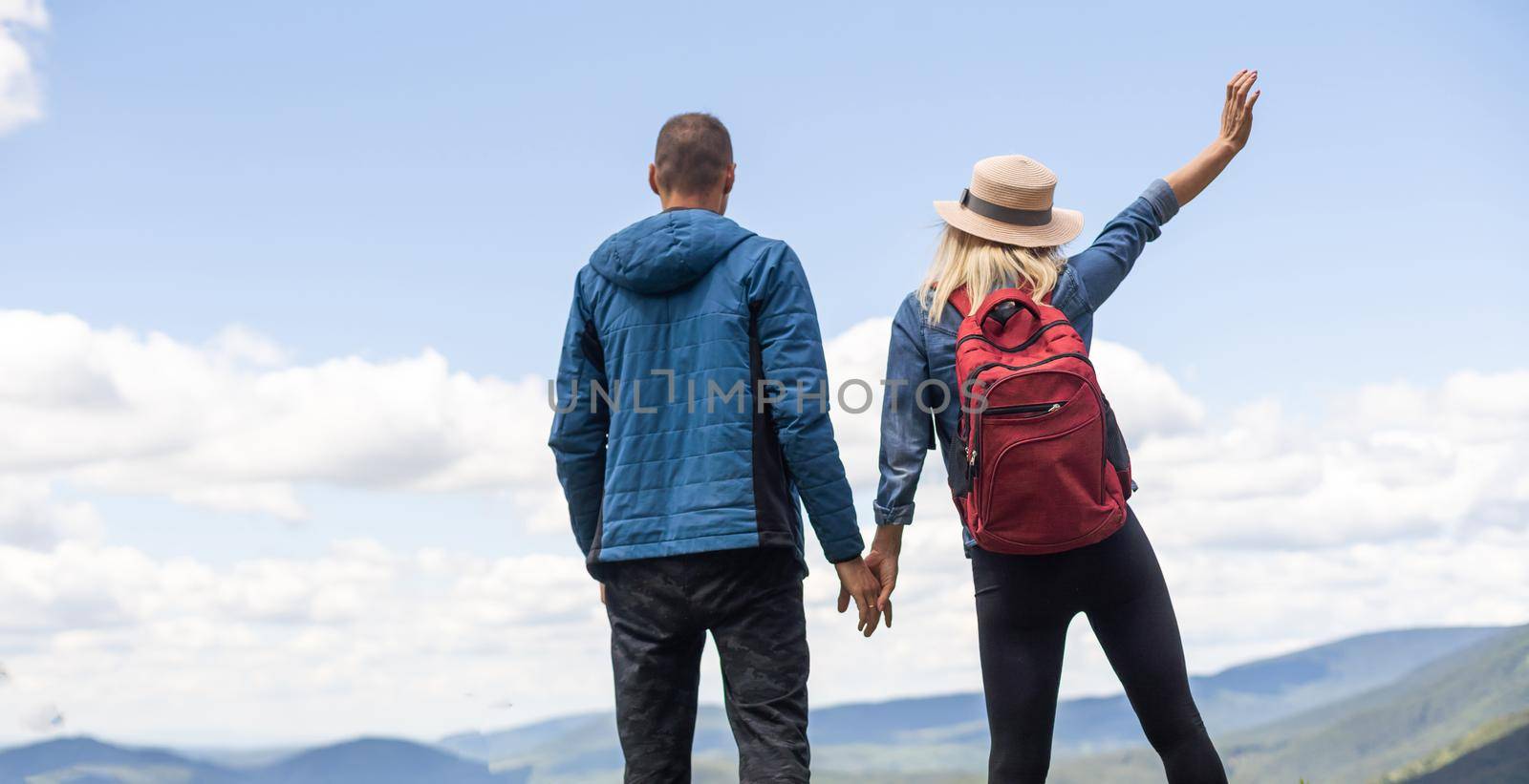 Rear view of hiking couple with backpack standing together on hill top enjoying beautiful landscape. Man and woman outdoors on hiking standing on a rock by Andelov13