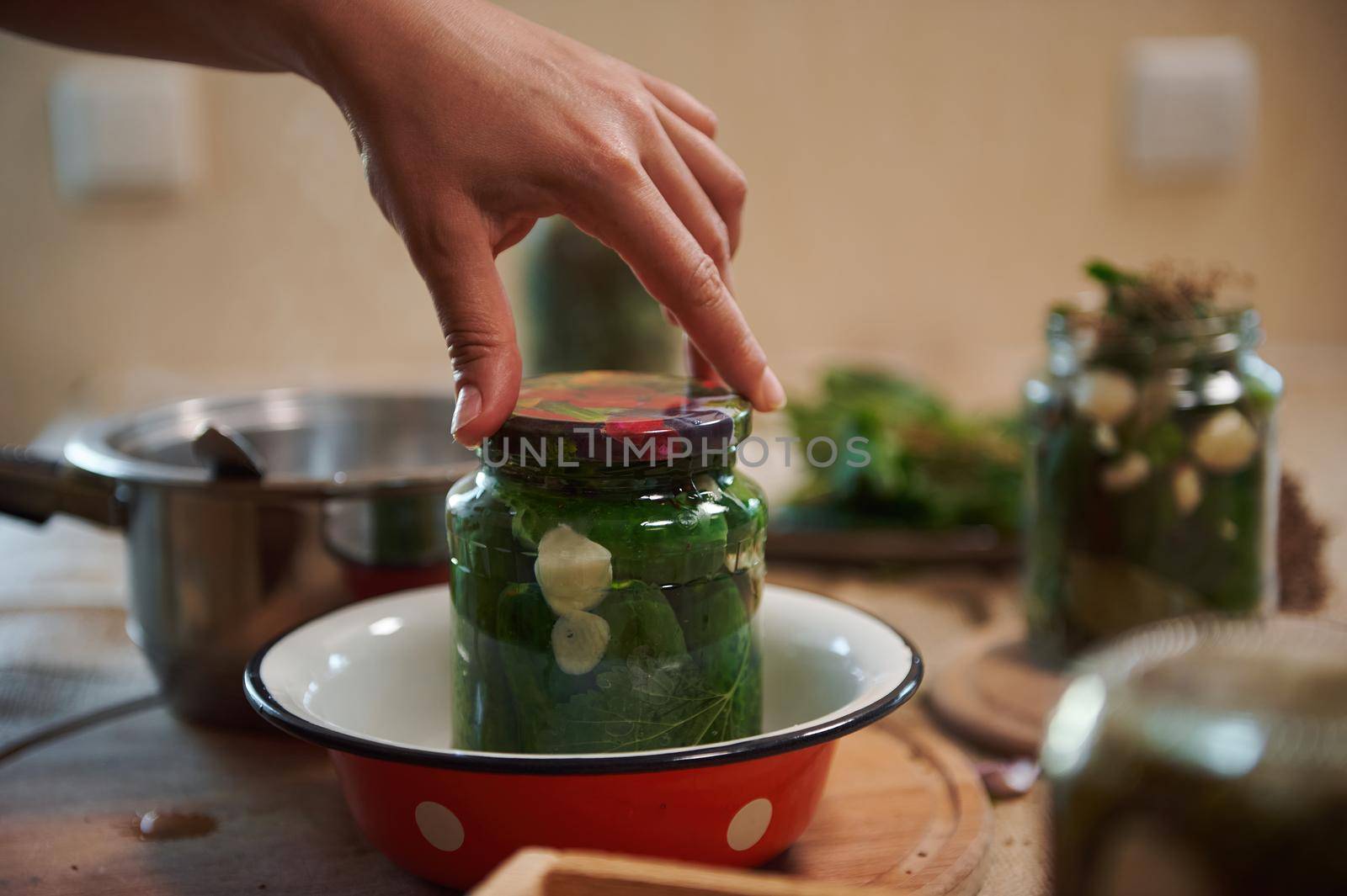 Details: Chef's hand putting a lid on a sterilized jar with cucumbers, pickled with fresh dill leaves, garlic cloves and fragrant culinary herbs