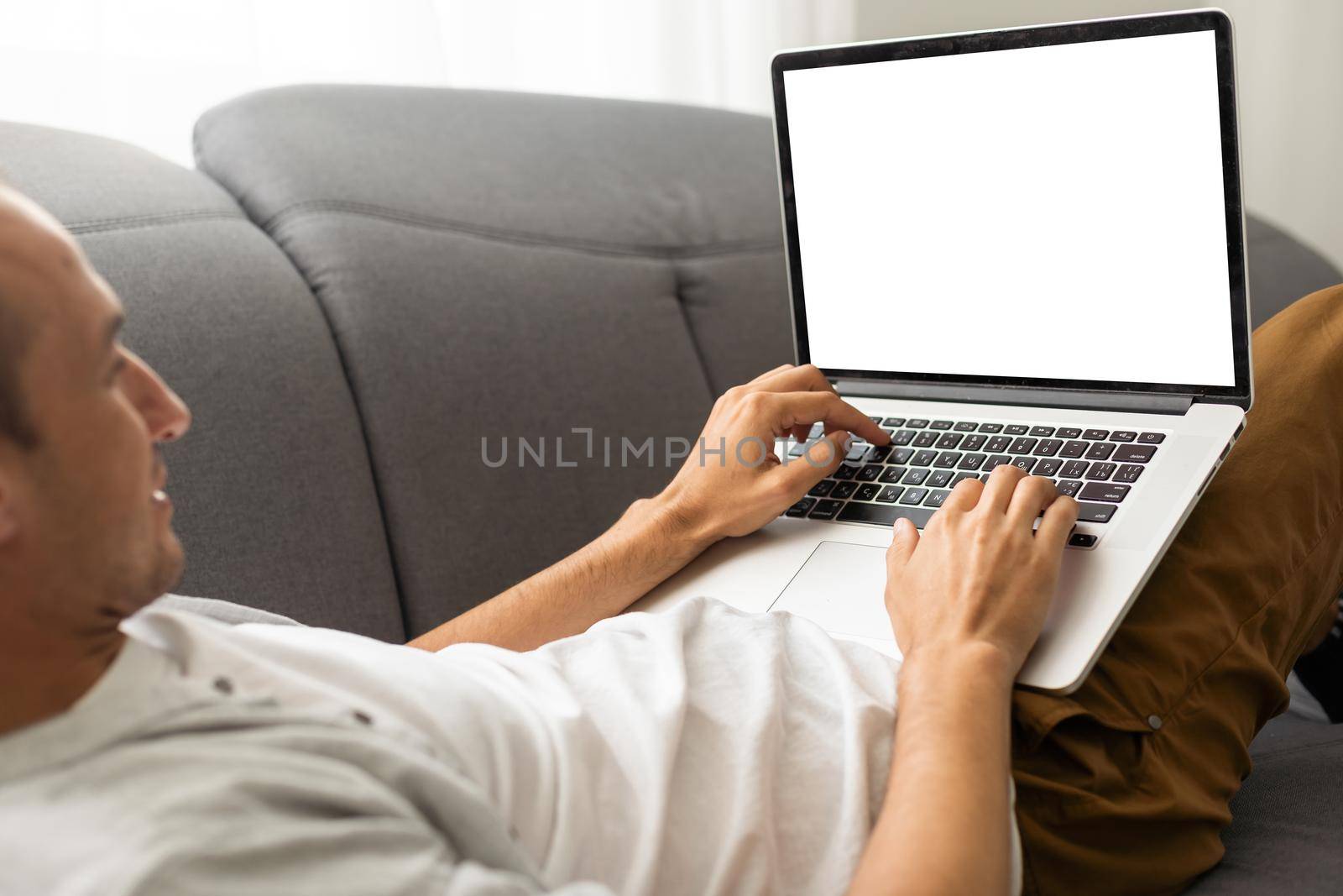 Young male tech user relaxing on sofa holding laptop computer mock up blank white screen. Man using modern notebook surfing internet, read news, distance online study work concept. Over shoulder view. by Andelov13