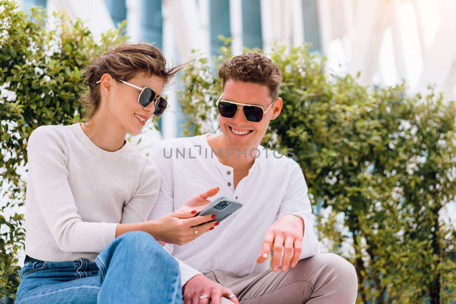 Modern Businessman showing something in the smartphone to his female assistant. Modern business young business people in sunglasses using smartphone smile sitting bench outdoor in office district