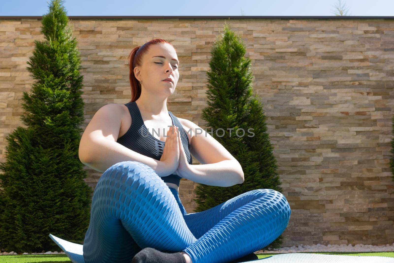 young woman with red hair, practising yoga in the garden in the open air, relaxation exercises, doing the lotus posture. concept of health and well-being. by CatPhotography