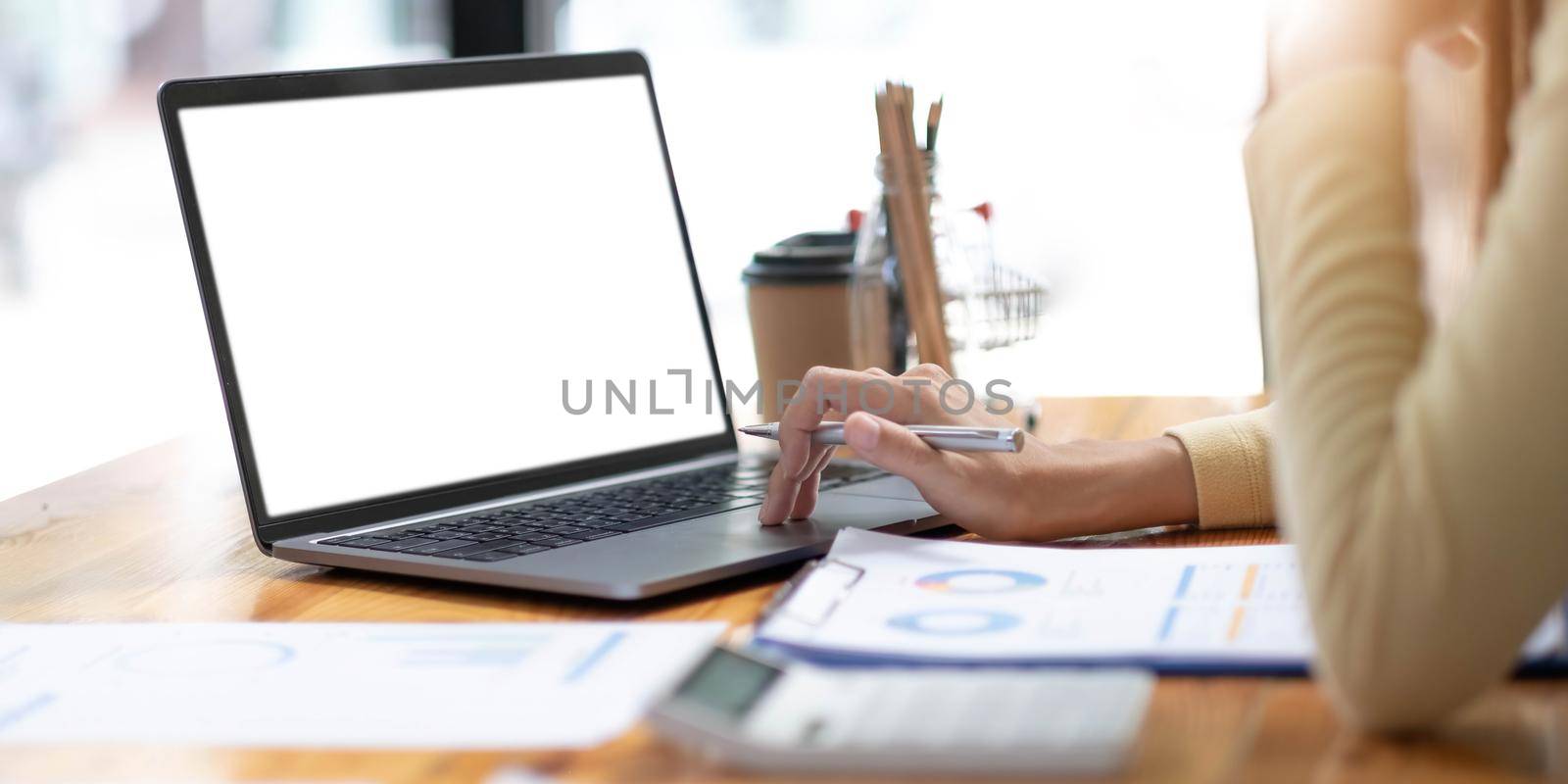 Mockup image of business woman using and typing on laptop with blank white screen and coffee cup on glass table in modern loft cafe.