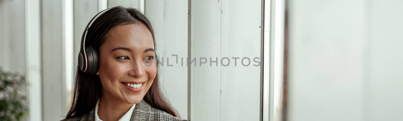 Asian Business woman listening music from phone in headphones standing near window in office